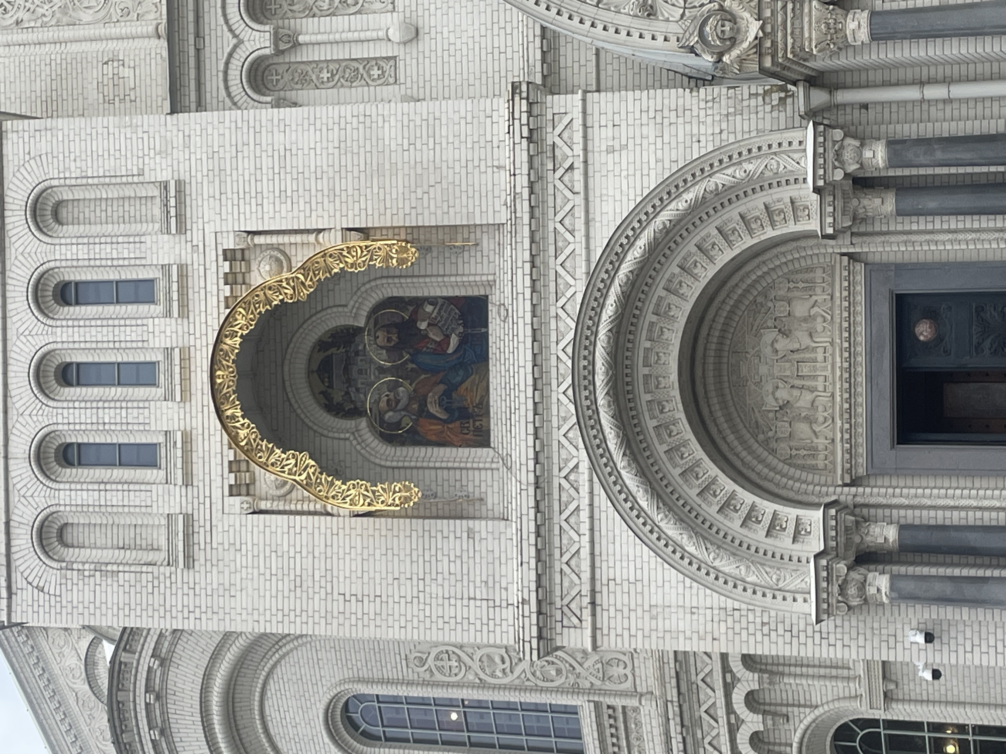 The image shows the exterior of the St. Nicholas Naval Cathedral in Kronstadt, Russia. The cathedral is constructed of white brick and features a large arched entrance with ornate carvings. Above the entrance, a golden archway frames a mosaic depicting two religious figures. The facade is adorned with decorative elements, including columns, arches, and windows. The lighting suggests it is daytime.