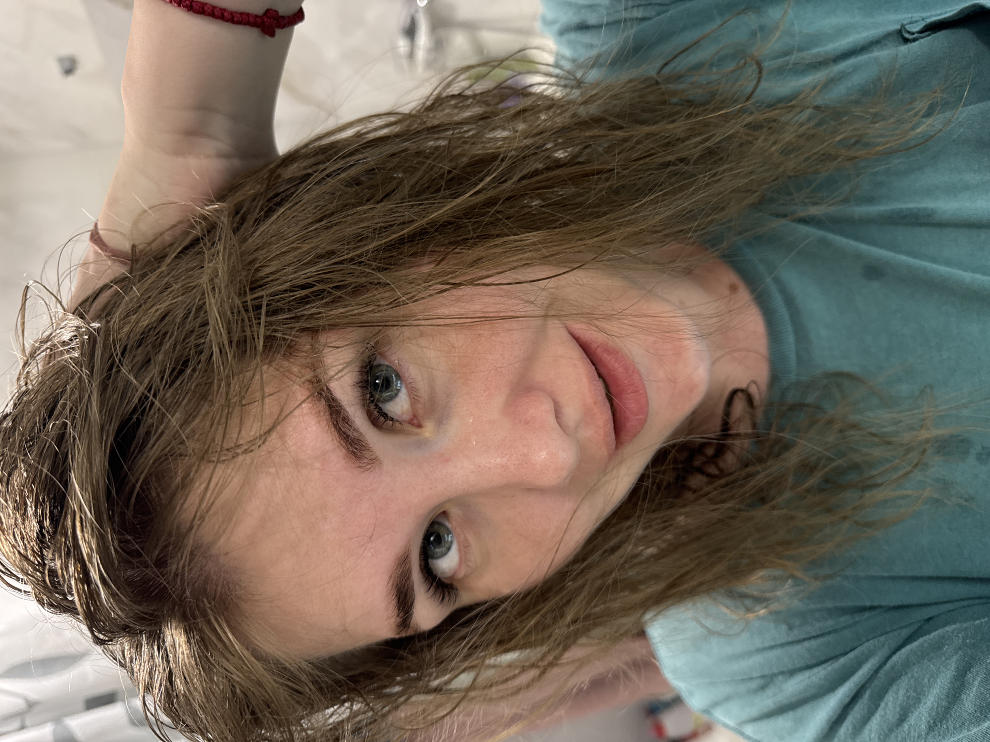 A close-up, rotated view features a young woman with light brown, wet hair and blue eyes. She is wearing a teal shirt and has two wristbands on her left wrist: one red and one pink. The background is blurry and appears to be a bathroom setting with white tiles. The location is not discernible as Ферма, Russia from the information within the picture.