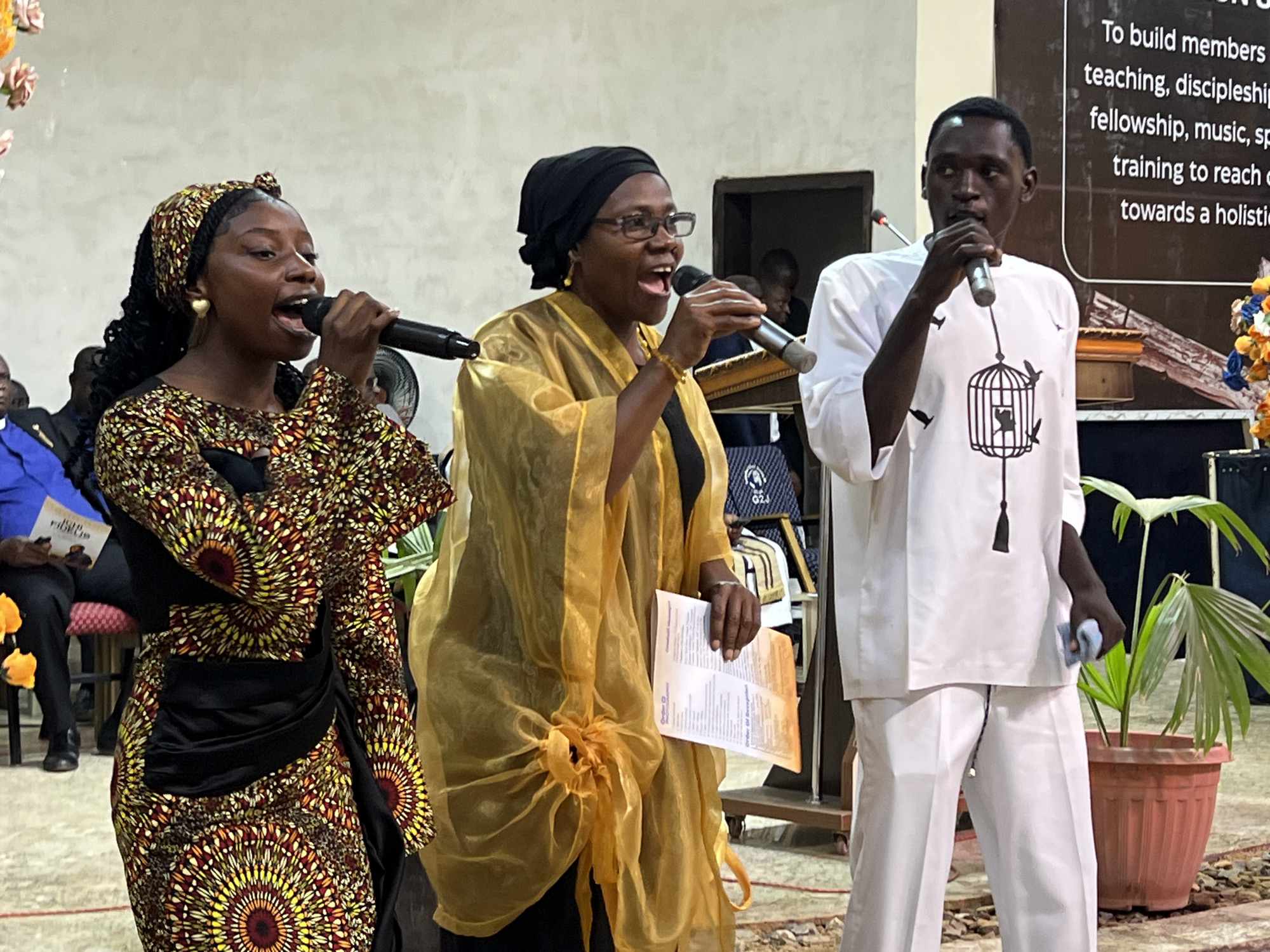 Three individuals are standing in what appears to be an indoor venue in Kabong, Nigeria, each holding a microphone and seemingly participating in a performance or service. The person on the left is a young woman wearing a vibrantly patterned dress with red, yellow, and black circular motifs, and a matching headscarf. She is singing into a microphone. Beside her, in the center, is an older woman with glasses, wearing a black headscarf and a flowing golden garment over dark clothing. She is also singing into a microphone and holding a pamphlet or paper. On the right, a man in a white tunic and trousers, adorned with a black birdcage design on his chest, holds a microphone in his right hand and a light blue cloth in his left. Behind them, a large brown sign with white text is visible, along with part of a doorway. The background suggests a community hall or event space, with plants in pots and seating areas. The lighting is bright and even.