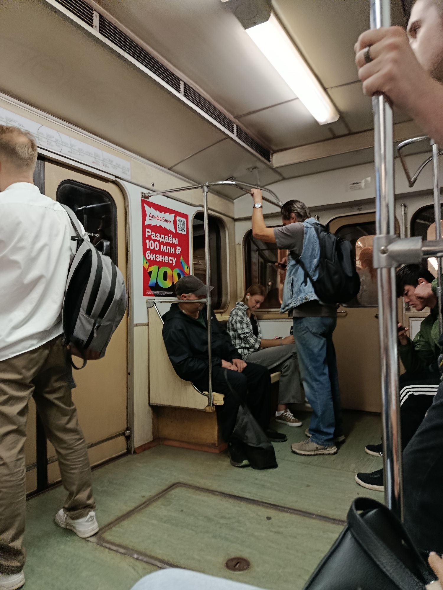 The image shows the interior of a subway car. In the foreground, a person in a white shirt and khaki pants stands facing away from the camera, wearing a grey backpack. To their left, a door is visible. Further inside the car, several passengers are seated and standing. A man in a dark jacket and cap is seated on a beige bench. To his right, a woman in a plaid shirt and jeans is seated, looking at her phone. A man in a denim vest and jeans stands holding onto a pole and looking at his phone. Another man, wearing a green jacket, is seated on the right side of the frame, also looking at his phone. The subway car has beige walls, a grey ceiling with fluorescent lighting, and a green floor. A red and white advertisement is visible on the wall. The overall scene depicts a typical commute on public transportation.