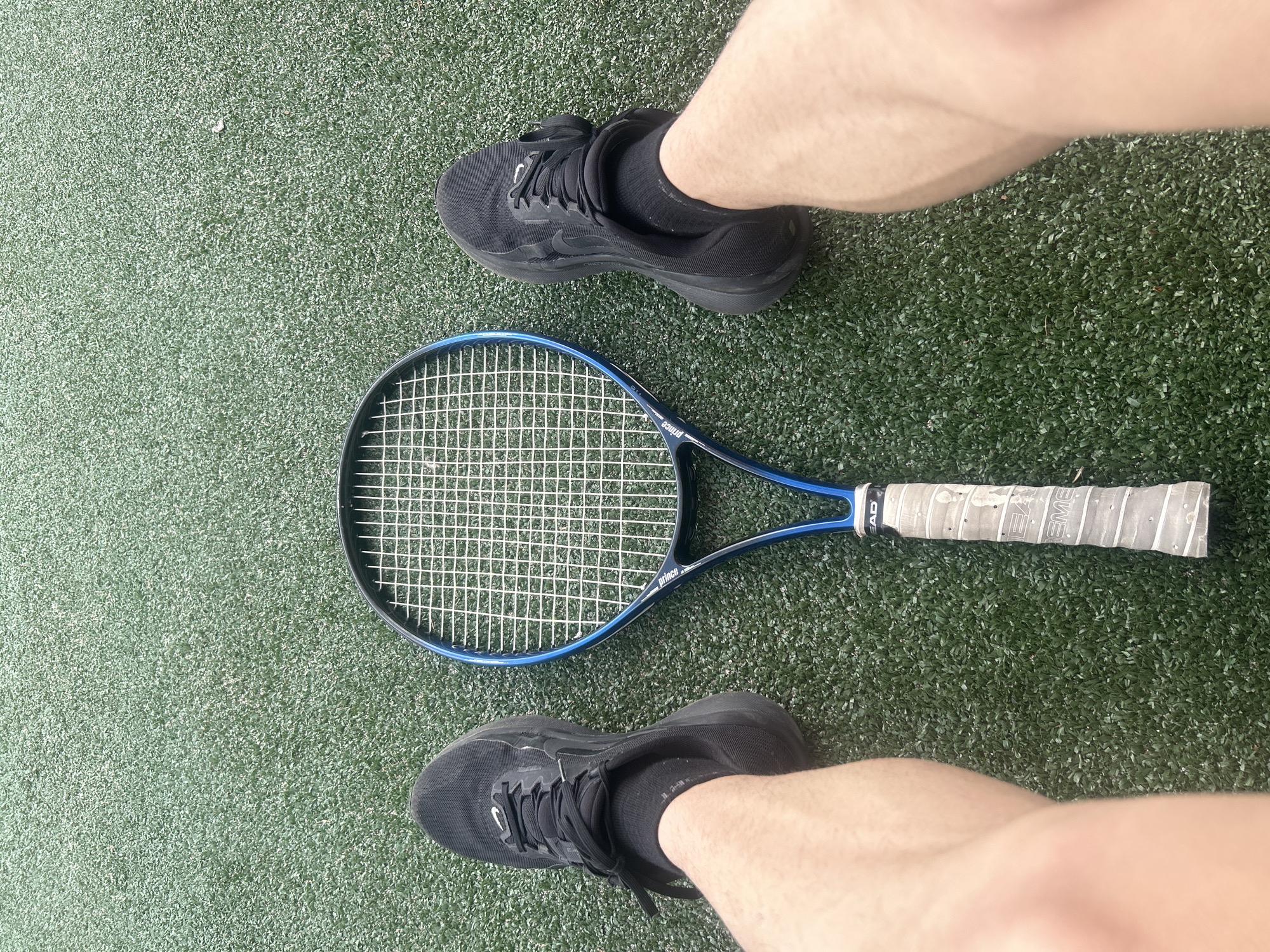 The image shows a tennis racket lying on a green surface, likely artificial grass or a tennis court. The racket is blue with a white grip. Two feet wearing black shoes and black socks are positioned on either side of the racket. The image is taken from a high angle, looking down at the scene. There is no information in the image to indicate it was taken in Veliko Tarnovo, Bulgaria.
