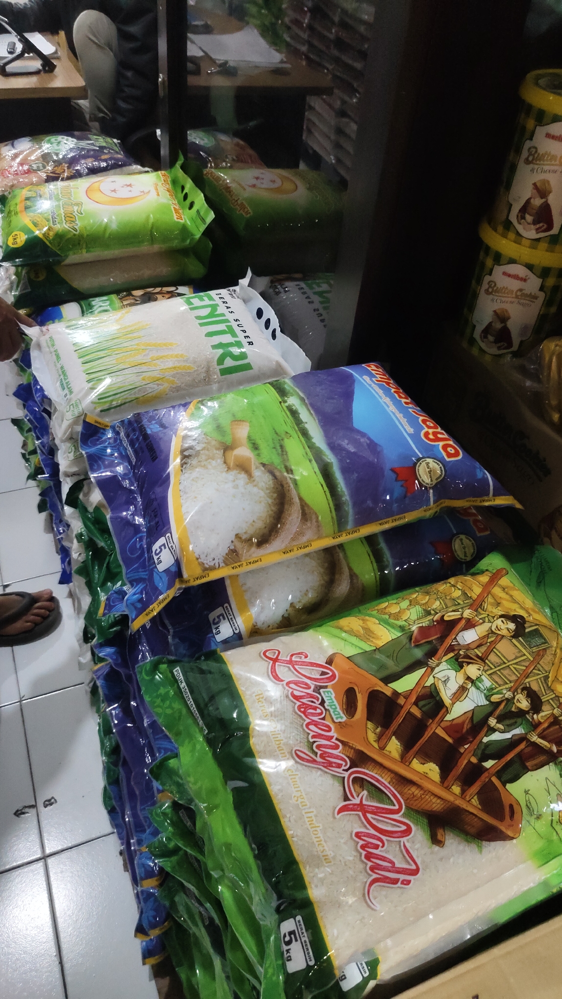 The image depicts a retail environment in Malang, Indonesia, showcasing stacks of rice sacks and canisters of cookies. The primary focus is on a variety of rice packaging, mostly in plastic bags, some with vibrant graphics. One prominent bag features a crescent moon and star motif on a green background, indicating a potential brand. Another bag displays an illustration of rice grains in a wooden bowl with a scoop. Further down, a bag with the branding "Empat Padi" is visible, adorned with an illustration of people in a boat. The rice sacks are stacked in a dense arrangement, suggesting a commercial setting like a grocery store or market. To the right, several yellow canisters with a checkered pattern and the label "merlin bak" and "Butter Cookies & Cheese" are stacked, suggesting a complementary product offering. A person's feet, wearing a sandal, are visible on the lower left, partially obscuring the bottom of the rice sacks. In the background, a person is partially visible, seated and looking towards a tablet or computer screen, with a desk and more merchandise in the background. The overall environment appears to be an indoor retail space with tiled flooring.