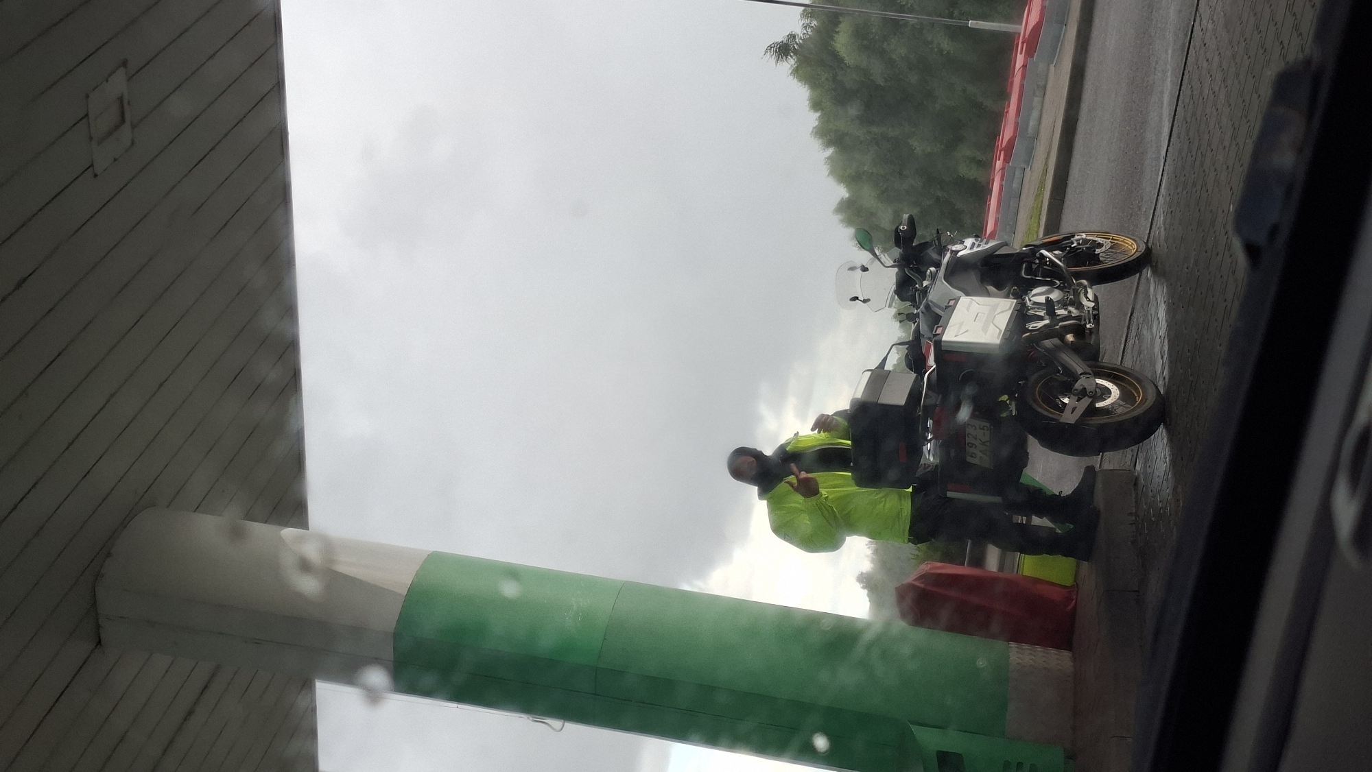 A person wearing a bright yellow raincoat and dark pants stands next to a motorcycle at a gas station. The person is holding a bottle and appears to be in the process of filling it. The motorcycle is a dual-sport model with saddlebags and a top case. The gas station has a distinctive green and white support pillar and a red barrier in the foreground. The ground is wet, suggesting recent rain. In the background, there are trees and a cloudy sky. The scene is viewed from inside a vehicle, with the car's dashboard visible in the bottom of the frame, indicating the photographer is a passenger. The license plate on the motorcycle reads "6923 AK-5". The setting is the Myadzyel Gas Station #2, located on the Mityunsky highway, 1.5 kilometers from Myadzyel, Belarus.