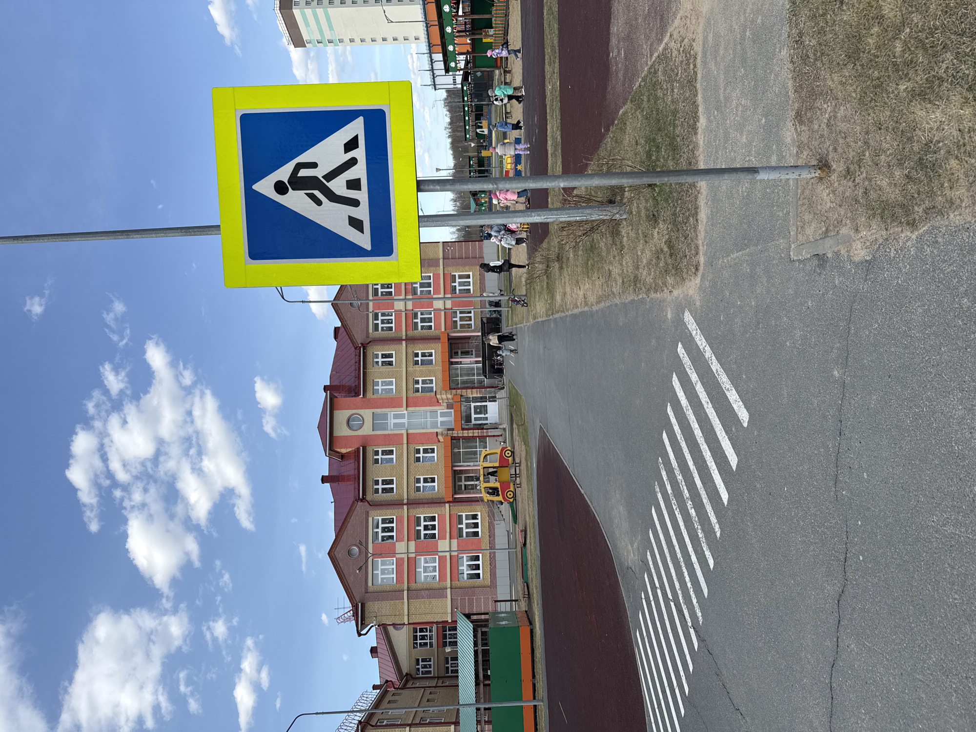 The image, taken during daytime under a clear blue sky with scattered white clouds, captures an outdoor scene in what appears to be a residential or educational area in Tobolsk, Russia. The primary subject is a pedestrian crossing sign, visible prominently on the left side of the frame. The sign features a yellow square border around a blue square, which contains a white triangular warning symbol with a black silhouette of a person walking. This sign is mounted on a metal pole.
Beneath the sign, and extending to the right, is an asphalt path or road section, with white diagonal striped markings indicating a pedestrian crossing. Adjacent to this asphalt, on the right, is a section of patchy grass.
In the mid-ground, a multi-story building with a red roof and facades featuring a mix of red/orange brickwork and lighter panels, along with numerous white-framed windows, stands out. In front of this building, there's a children's playground area with green and dark red/brown soft surfacing. Several children and some adults are scattered throughout this playground, engaged in various activities. Playground equipment, including what looks like a yellow and red car-shaped structure, is visible. In the background, another taller, light-colored residential building is partially visible.
The overall scene depicts a typical neighborhood setting, possibly near a school or kindergarten, with designated areas for both pedestrians and children's play. The bright sunlight suggests fair weather.
