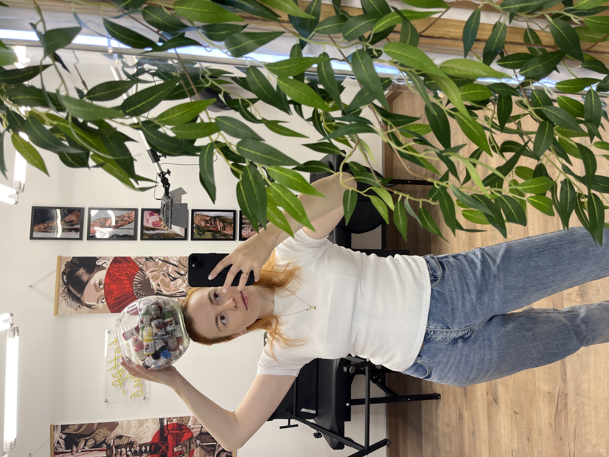 A young woman with auburn hair and freckles is taking a mirror selfie in what appears to be a tattoo parlor. She is wearing a white ribbed t-shirt and blue jeans. On her head, she is balancing a clear glass bowl filled with small bottles of tattoo ink. In her right hand, she holds a black smartphone, capturing the reflection. In the background, there is a black tattoo chair, artwork on the walls, and a large artificial plant with green leaves. The floor is made of light-colored wood. The scene does not provide any specific indicators to place it in the city of Chaikovsky, Russia.