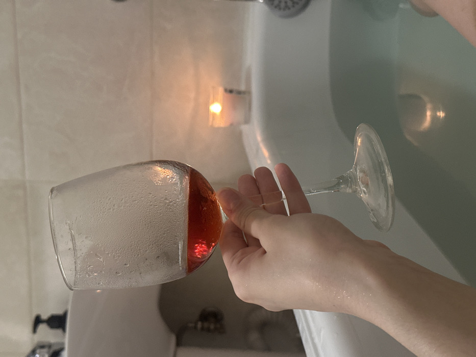 The image depicts a human hand holding a clear, stemmed wine glass containing a reddish-amber liquid, approximately one-quarter full. The exterior of the glass bowl is covered in visible condensation droplets. The hand, which appears wet, grasps the stem of the glass. The scene is set in a dimly lit bathroom in Khimki, Russia. A white bathtub, partially filled with clear water, occupies the lower right of the frame. A single lit tealight candle in a clear holder rests on the edge of the bathtub, casting a warm, orange glow that reflects on the water's surface and the glass. A portion of a chrome shower head is visible on the right edge of the tub. In the upper left background, part of a white ceramic sink or basin is visible, with a black pump-top dispenser on its edge. The walls in the background are light-colored, possibly tiled.