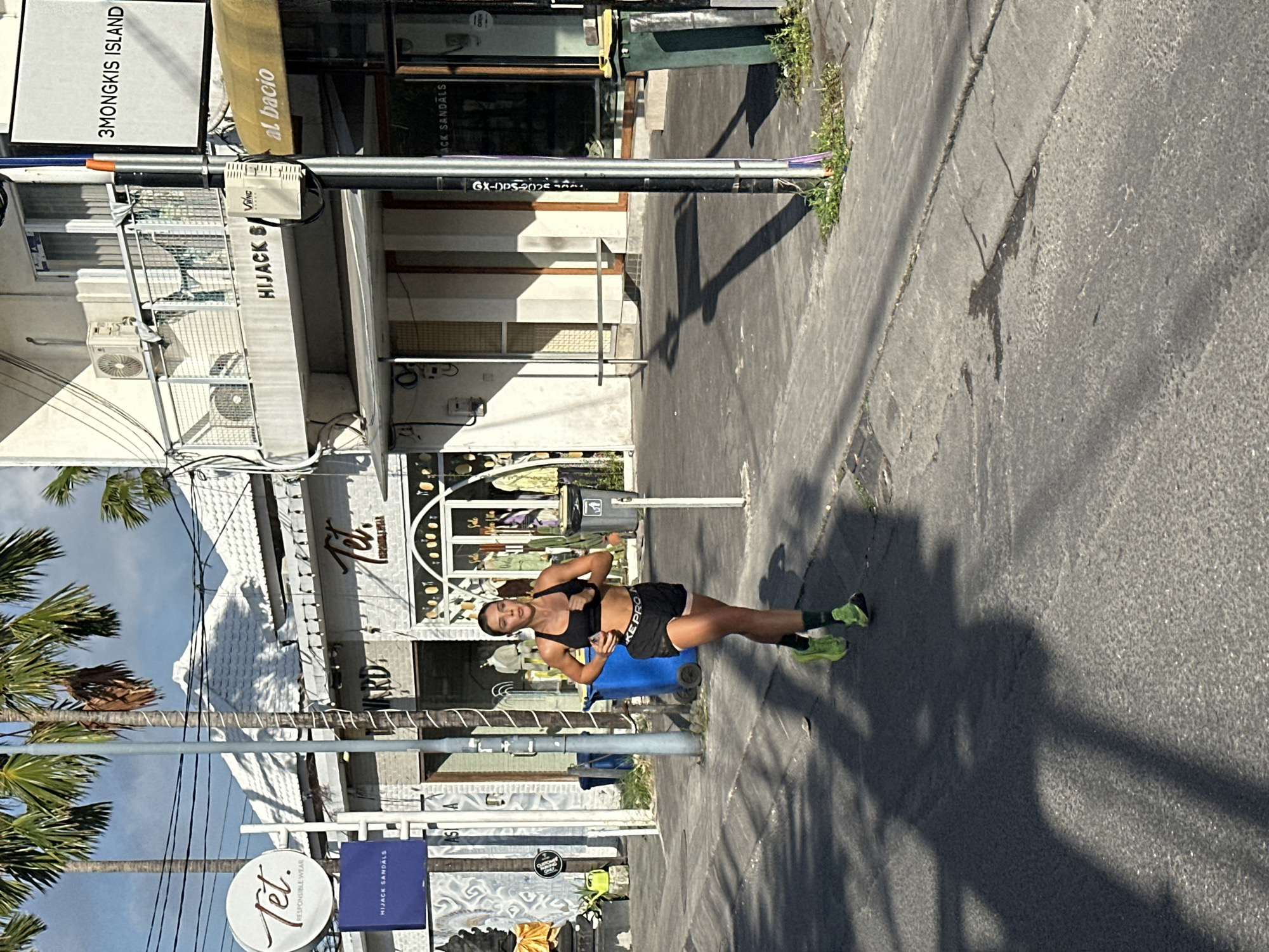 In North Kuta, Indonesia, a woman is captured mid-stride while jogging on a paved sidewalk adjacent to a street. She is wearing athletic attire, consisting of a black sports bra and black shorts with white lettering, and lime green running shoes with dark socks. Her hair is pulled back, and she appears to be focused ahead. The background reveals a commercial street with shops, including one with a sign that reads "Tet. Responsible Wear" and another with "Hijack Sandals" visible on a blue sign. Palm trees line the street, and shadows from them and possibly overhead wires are cast across the pavement. A street lamp is also present. The scene depicts an outdoor urban environment with signs of commerce and vegetation.