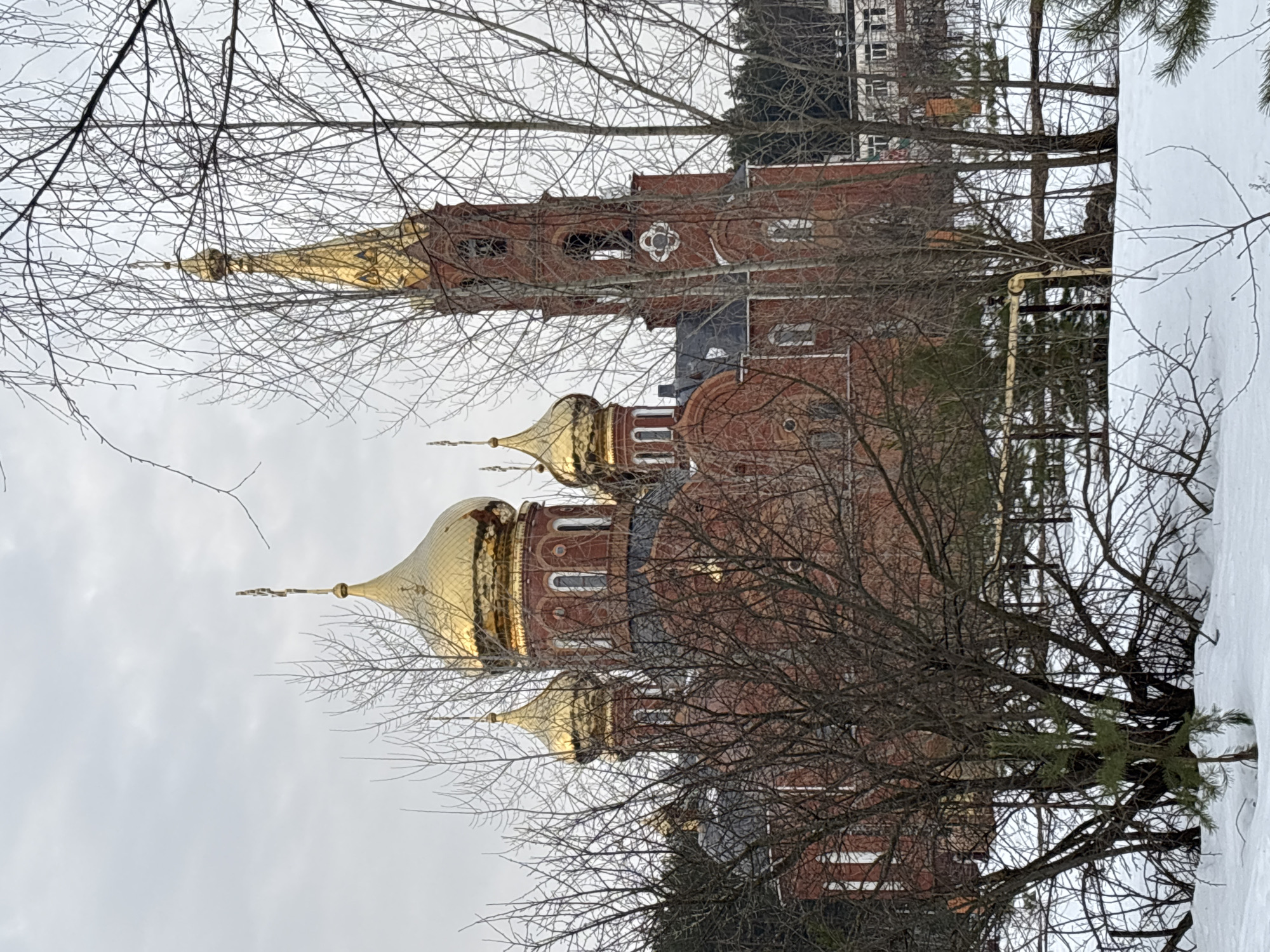 The scene depicts the Church of St. George the Victorious, located at ulitsa Stroitelnaya, 13, Chaikovsky, Perm Krai, Russia, under an overcast, light gray sky. The prominent structure is a multi-domed Orthodox church constructed of red brick. At least five golden onion domes, each surmounted by an Orthodox cross, are visible atop the main body of the church. A tall, slender bell tower, also of red brick, stands on the right side of the church, featuring arched bell openings, a white decorative circular element below them, and a golden spire topped with an Orthodox cross. The ground in the foreground is covered with a layer of white snow. Numerous bare deciduous trees and several small evergreen saplings are present in the foreground, partially obscuring the lower and mid-sections of the church. A horizontal yellow pipeline is discernible in the mid-ground, running behind some of the bare trees. In the distant background, a dark treeline and indistinct, light-colored buildings are visible. No human subjects are present within the frame.