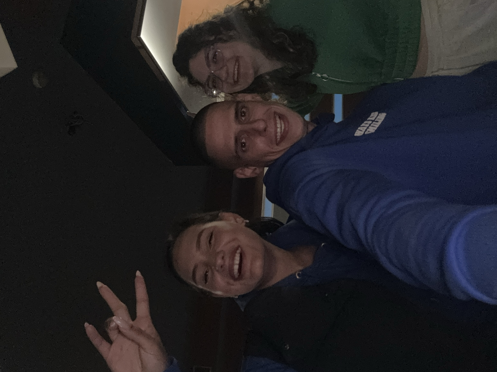 Three people are visible in the image. One woman is wearing glasses and a green top. A man with a shaved head is wearing a blue hoodie. Another woman is wearing a blue jacket and making a hand gesture. The environment is dark and appears to be indoors. The location is not discernible as Sofia, Bulgaria.