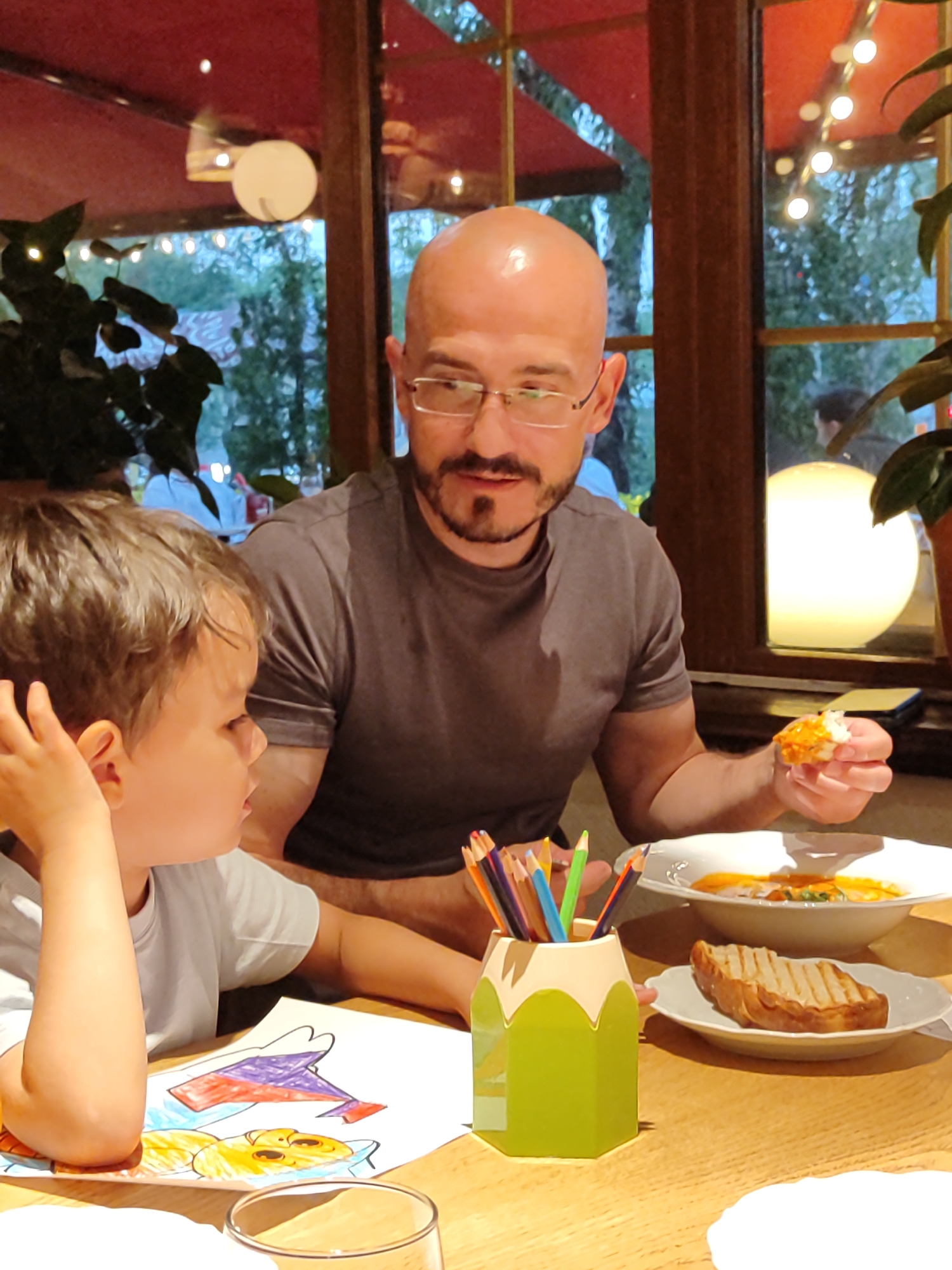 A man and a young boy are seated at a wooden table in a restaurant. The man, bald and wearing glasses, is looking down and holding a piece of food to his mouth. The boy, with brown hair, is looking at the man and has his hand near his ear. In front of the boy is a drawing with colored pencils in a green pencil-shaped holder. On the table, there are two plates with food, one with soup and the other with toast. Behind them, a large window offers a view of trees and lights. The restaurant has red ceiling panels and warm lighting.