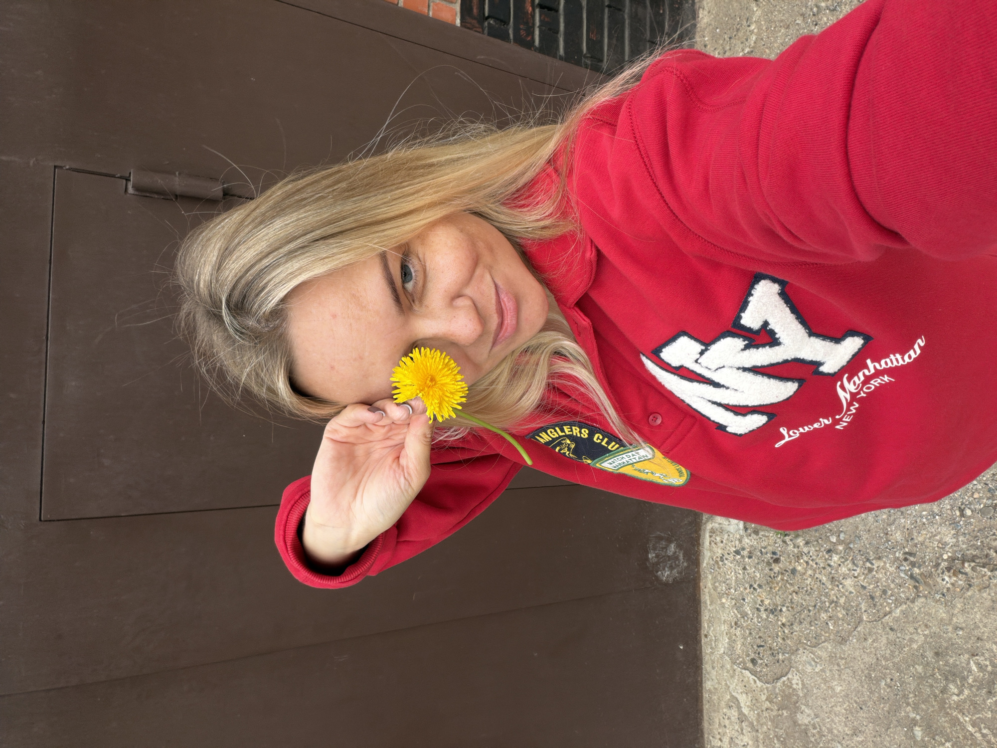 A blonde woman with blue eyes holds a yellow dandelion in front of her right eye. She is wearing a red sweatshirt with "NY Lower Manhattan New York" printed on it. The background appears to be a brown garage door and a brick wall. The image is taken outdoors in an urban setting.