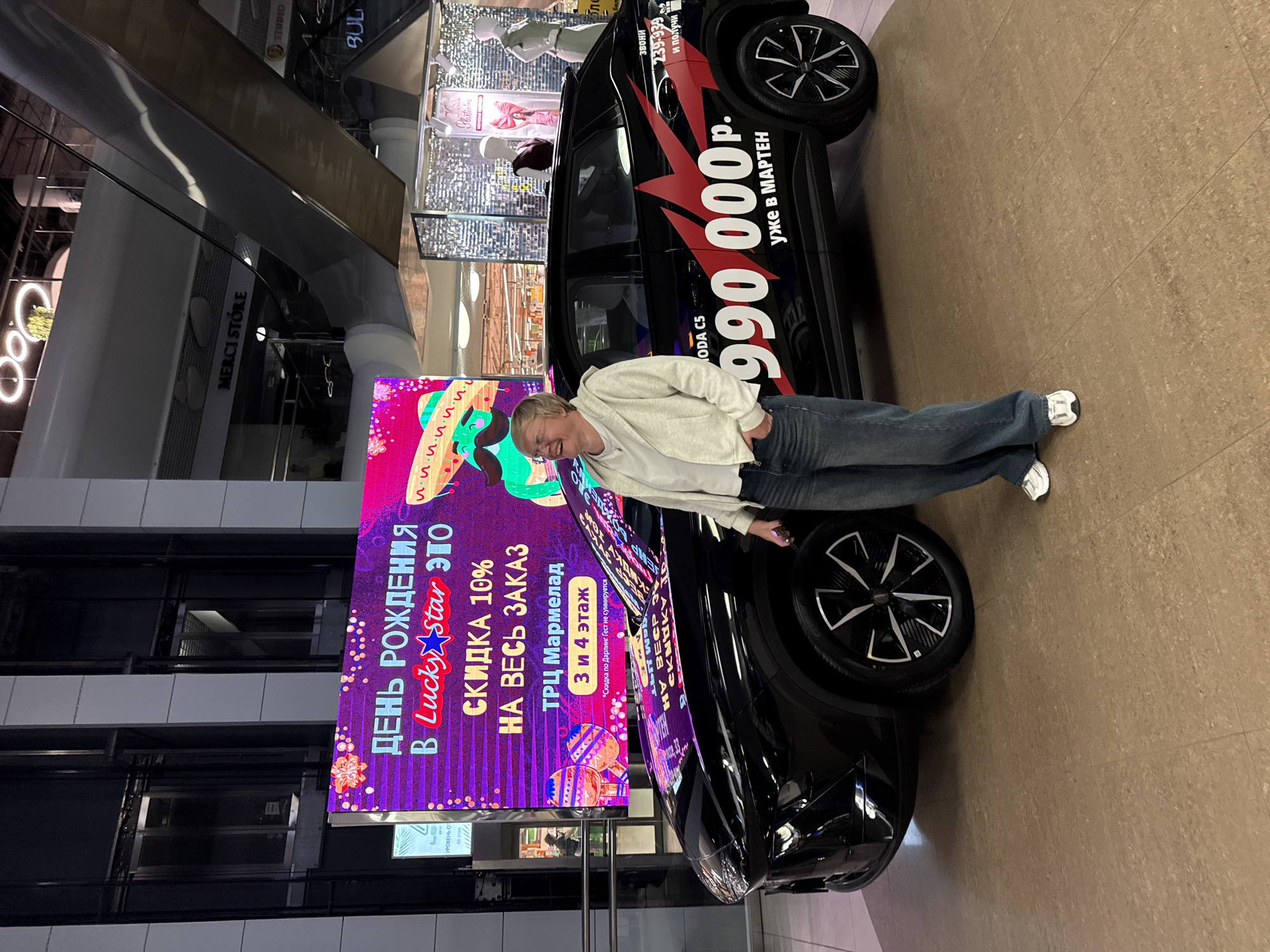 A woman stands in front of a black car with promotional graphics in a shopping mall. The woman is wearing glasses, a light-colored hooded jacket, and blue jeans. She is smiling and looking to her right, with her left hand in her jeans pocket. The car has large red graphics on its side, including the text "990 000 p." and "уже в МАРТЕН." A large digital screen behind the car displays promotional text in Russian, including "ДЕНЬ РОЖДЕНИЯ В Lucky Star ЭТО СКИДКА 10% НА ВЕСЬ ЗАКАЗ" and "ТРЦ Мармелад 3 и 4 этаж". The mall environment is visible in the background, with escalators and storefronts. The floor is tiled.