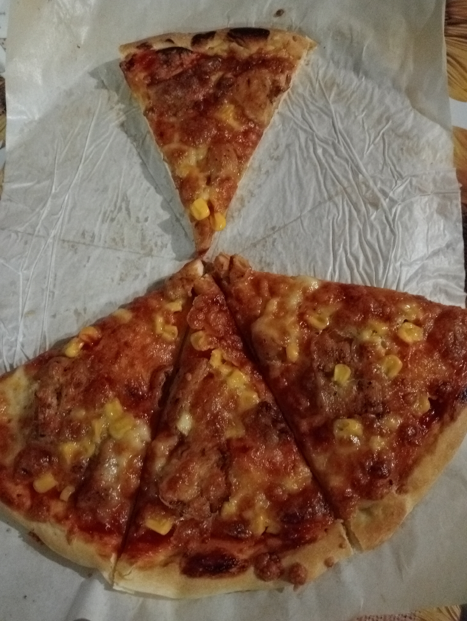 An overhead, medium shot captures four slices of pizza arranged on a crumpled white paper liner, which is itself placed on a patterned placemat. A single, triangular slice sits at the top, positioned vertically with its crust facing downwards. Below this, three more slices are arranged in a fan-like pattern, overlapping each other. The pizza appears to be topped with tomato sauce, melted cheese, and visible kernels of corn. The crust of all slices is golden brown. The lighting is dim, and the image has a grainy quality. The scene appears to be an indoor setting.