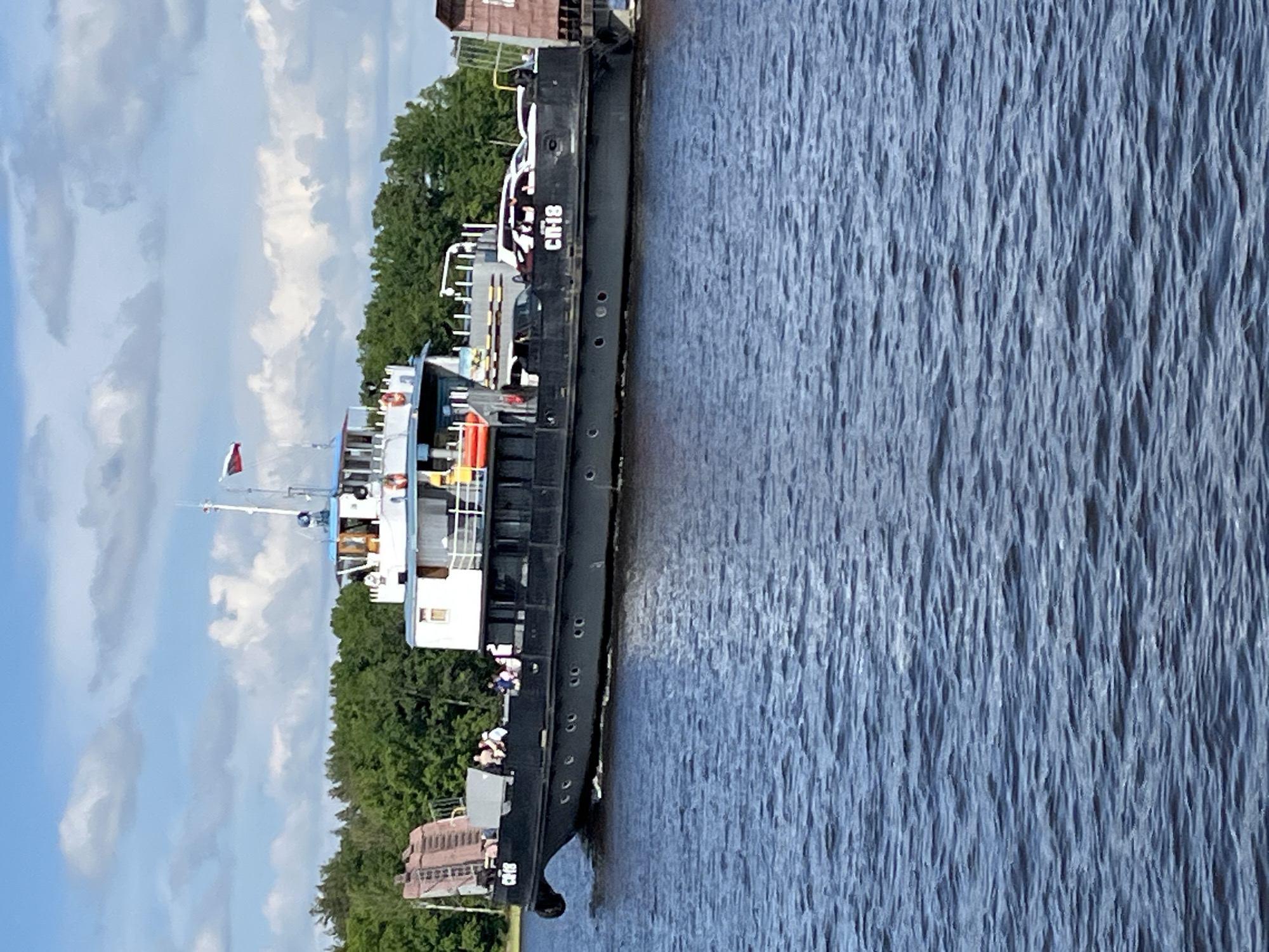 The image depicts a passenger and car ferry actively traversing a body of water under a bright, partly cloudy sky in the vicinity of Kazan, Russia.
The ferry, identified by the white Cyrillic alphanumeric designation "СП-18" (SP-18) on its dark grey or black hull, moves from left to right. Its superstructure is predominantly white with a light blue upper deck and pilot house roof. A Russian flag (horizontal white, blue, and red stripes) flies from a mast above the pilot house. Visible on the port side of the superstructure are white railings and several orange life preservers.
On the ferry's main deck, at least three vehicles are visible: a prominent white SUV (possibly a Toyota Land Cruiser or similar model) parked facing forward on the right side, a dark-colored sedan partially visible behind it, and a third white car further back. The ferry's bow features a large, dark brown, multi-sectioned vehicle ramp in a raised position.
Approximately eight to ten individuals are visible on the open deck areas of the ferry's superstructure, most standing and casually observing their surroundings. They appear to be passengers, dressed in light, casual clothing appropriate for warm weather.
The water is a deep blue, exhibiting numerous ripples and small waves, indicating movement from either wind or the ferry's wake. In the distant background, a dense and vibrant line of green trees, a mix of deciduous and coniferous species, forms the shoreline. The sky above is a light blue with scattered, fluffy white cumulus clouds. The lighting suggests bright daylight conditions.