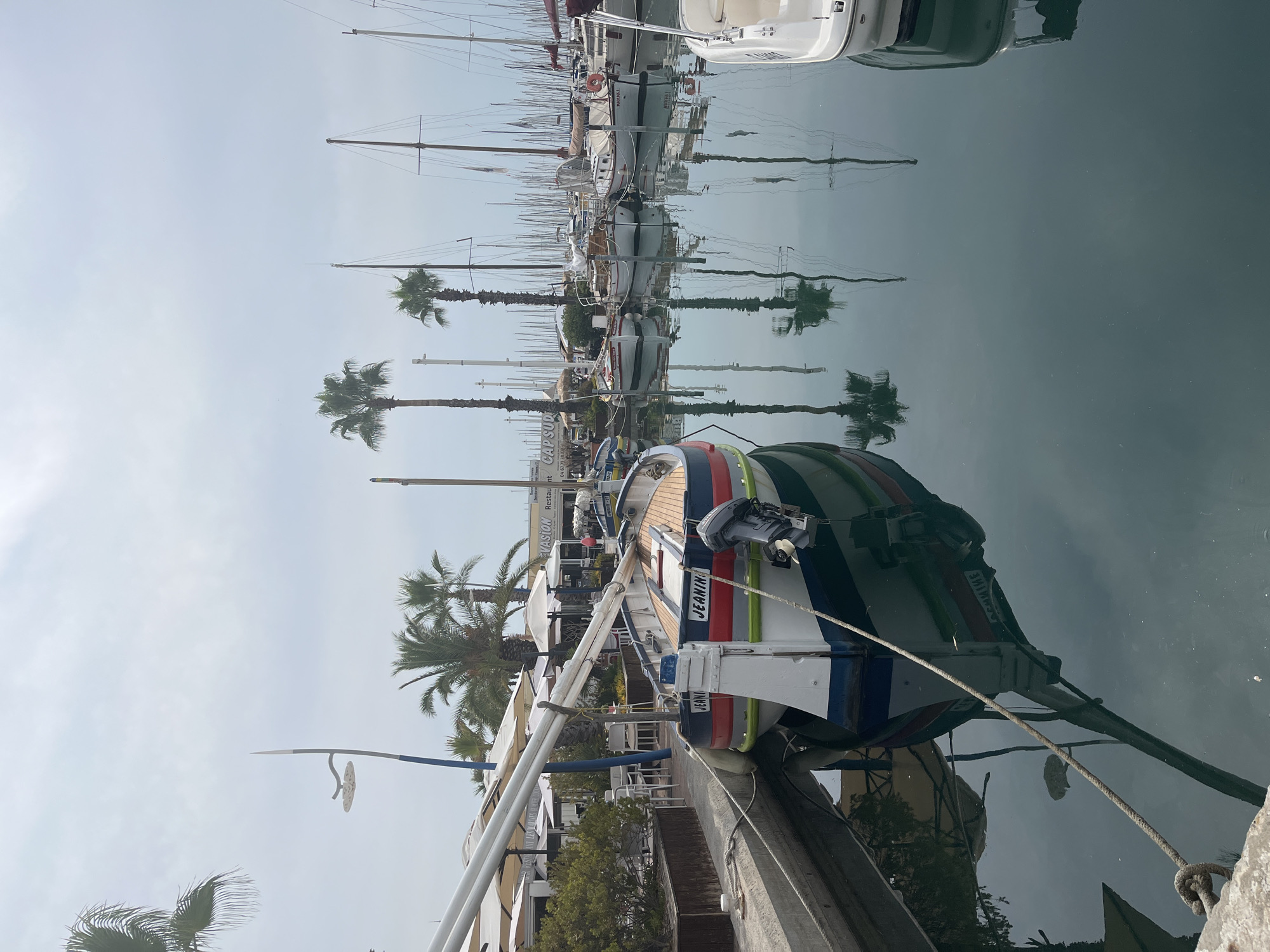 This is a photograph of a marina in Palavas-les-Flots, France, on a cloudy day. The foreground features a small sailboat tied to a dock, its name, "JEANIN," clearly visible on the stern. The boat is painted with red, white, and blue stripes along its hull. An outboard motor is attached to the back of the boat. The water in the marina is calm and reflects the boats, palm trees, and masts of other sailboats in the background. Several other boats are moored in the marina, including some white sailboats with their masts visible. Palm trees line the waterfront, and beyond them, buildings with white umbrellas suggest a waterfront restaurant or cafe. The sky is overcast and a pale blue.
