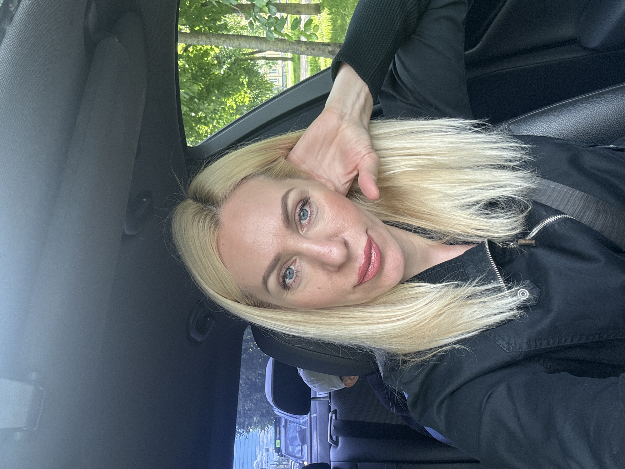 A blonde woman with blue eyes and pink lipstick is sitting in the passenger seat of a car, taking a selfie. She is wearing a black jacket and resting her hand on her cheek. In the background, through the car window, trees and buildings are visible. A child wearing a baseball cap is seated in the back of the car. The scene appears to be on a street in Saint Petersburg, Russia, with trees and buildings characteristic of the city visible outside the vehicle.