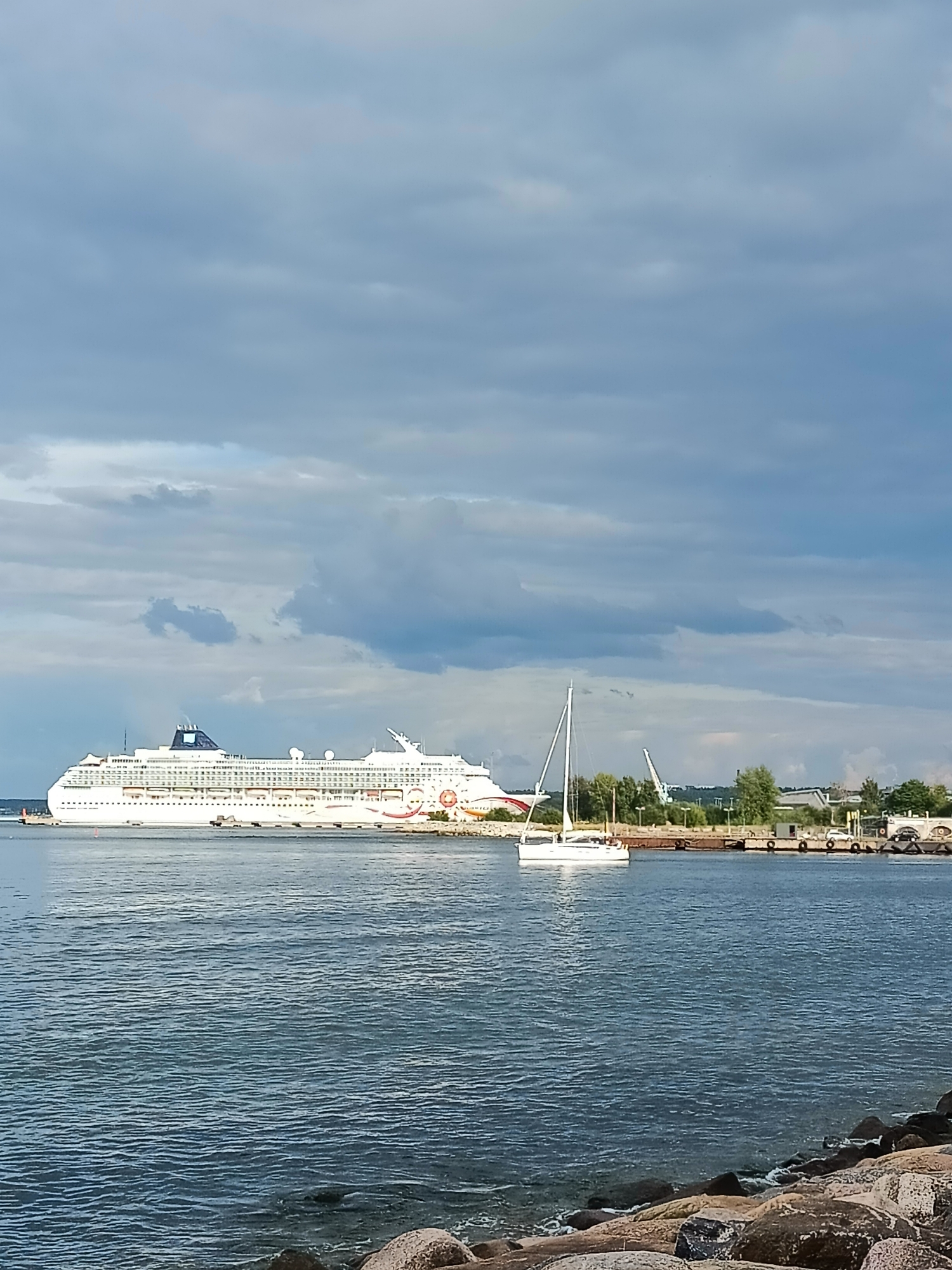 A large white cruise ship is docked at a port in Tallinn, Estonia. A white sailboat is sailing in the foreground, to the right of the cruise ship. The water is blue and calm, with small waves. The sky is cloudy and gray. In the background, there are buildings and trees. The scene appears to be a sunny day, despite the cloudy sky.