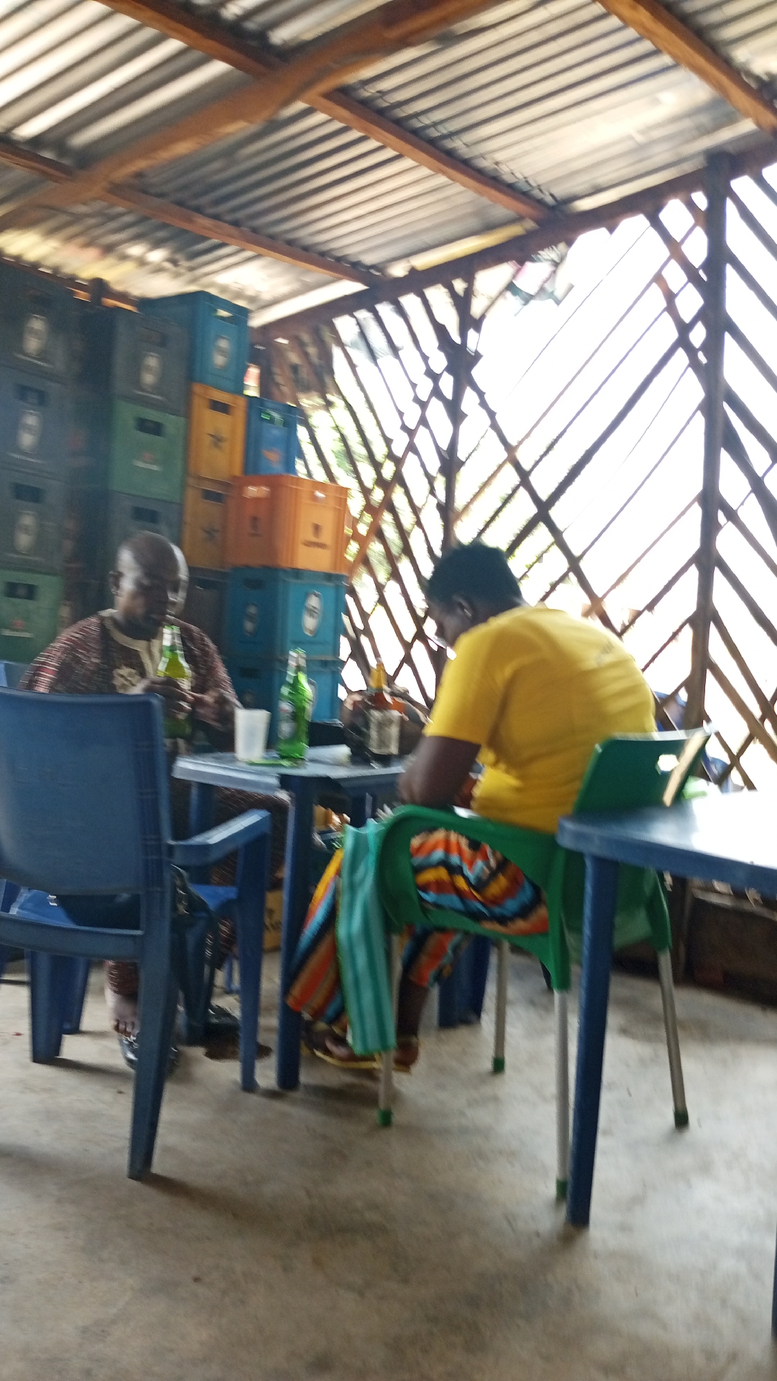 The image captures a scene in Zango-ndaji, Nigeria, where two individuals are seated at separate tables within an open-air structure. One person, dressed in a patterned brown shirt, sits at a blue plastic table, holding a green bottle, likely a beer. A stack of beverage crates in shades of green, blue, and orange is visible behind them. Across from them, another person wearing a yellow t-shirt and colorful patterned pants sits at a green plastic chair at a smaller table with a green cloth draped over the back. A green table is near them. The structure has a corrugated metal roof and lattice-like walls. The floor appears to be concrete.