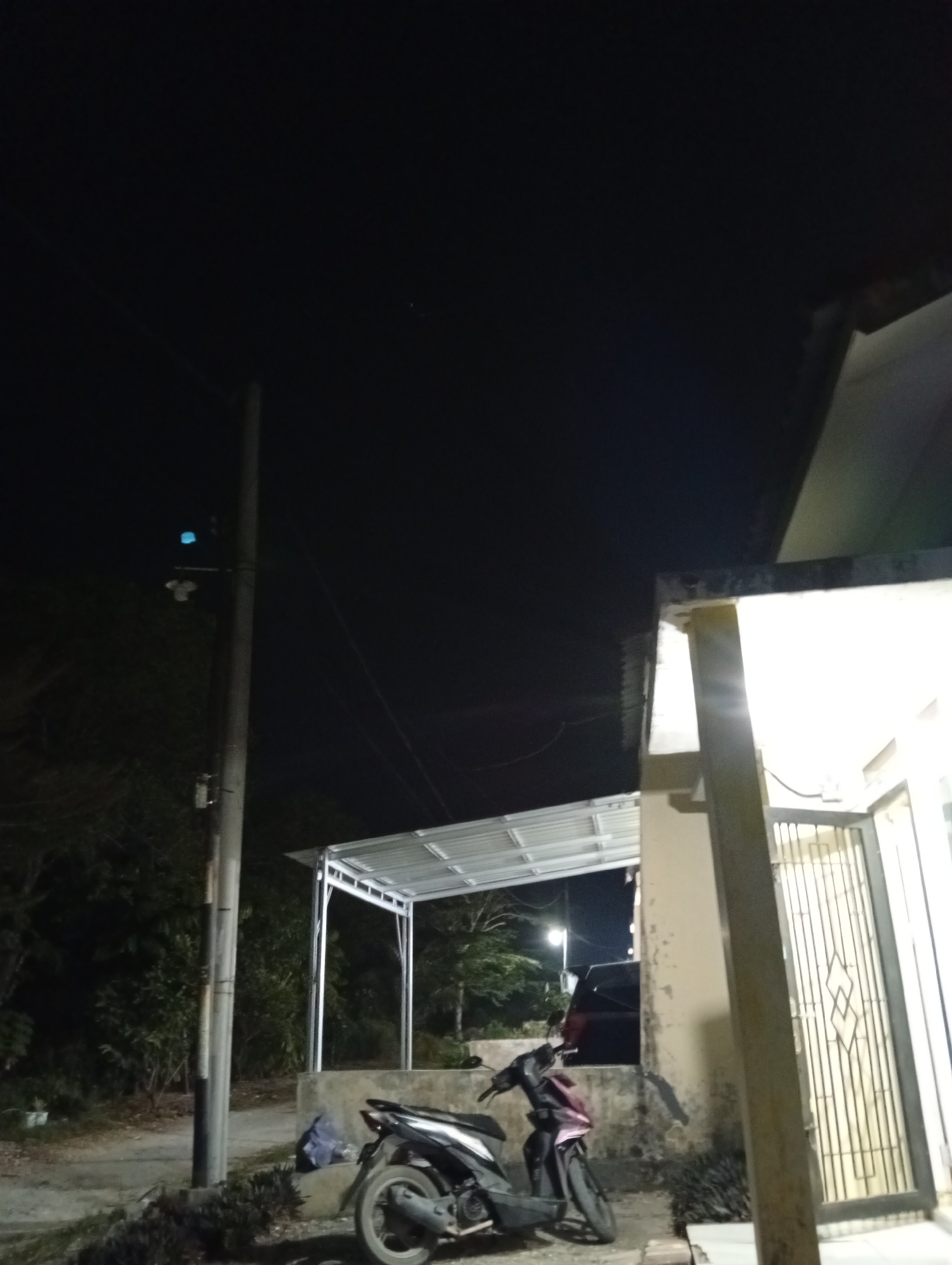The image depicts an outdoor scene at night, likely in a residential area of Kualu, Indonesia. In the foreground, a dark-colored motorcycle is parked on a paved surface. To the right of the motorcycle, a building with a closed metal gate and an illuminated window is visible. A concrete structure with a metal roof, possibly a carport or shelter, is situated behind the motorcycle. A utility pole with some attached equipment stands to the left of the motorcycle, with vegetation visible in the background. The sky is dark, indicating nighttime. No people are present in the image.