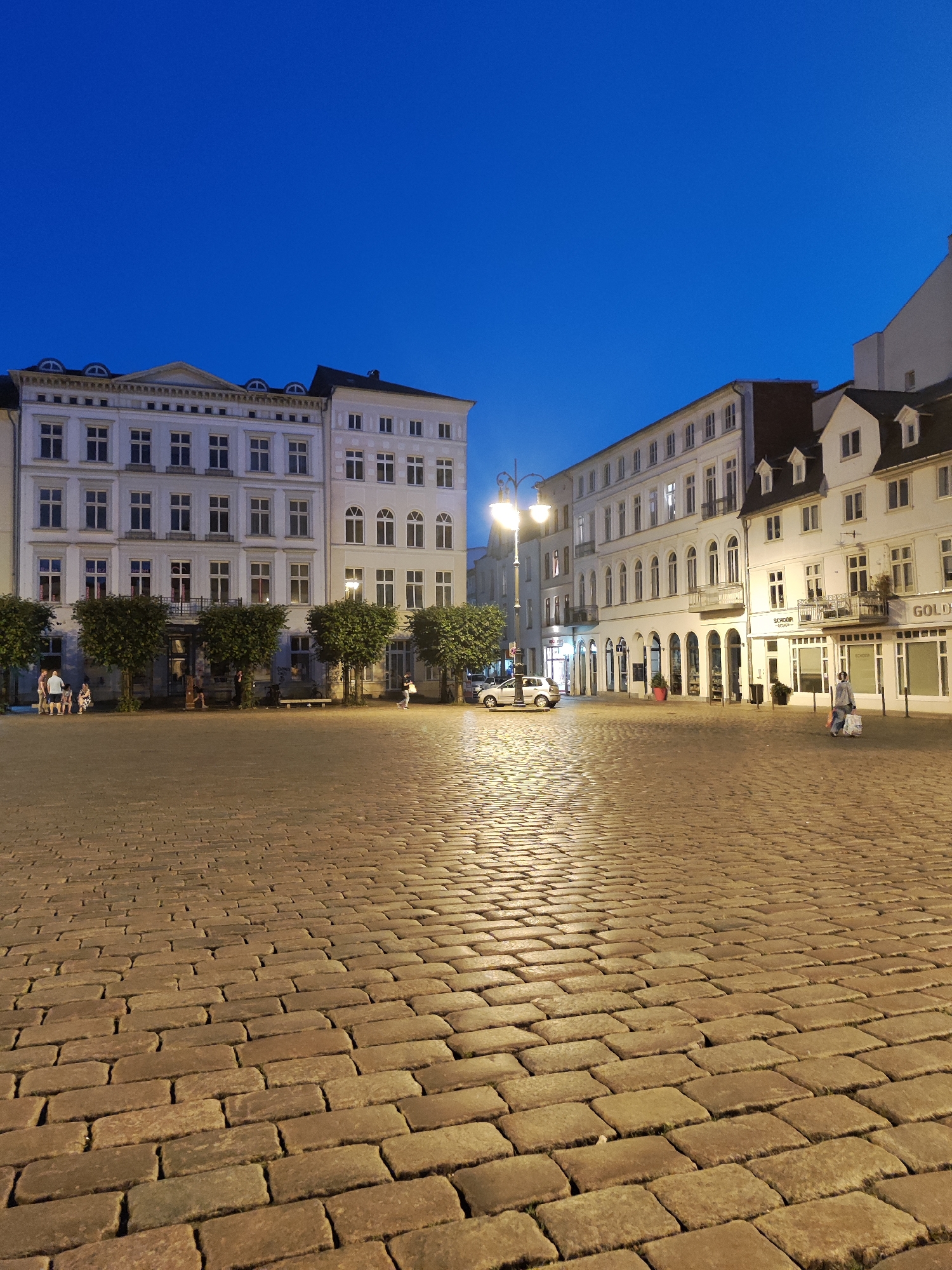 This is a medium-angle, vertical, eye-level shot of a cobblestone plaza at dusk in front of several European-style buildings. The sky is a deep, clear blue, indicating the transition from daylight to night. The foreground is dominated by a textured cobblestone ground, illuminated by the warm glow of streetlights that are visible in the middle ground.
Along the middle ground, a row of light-colored buildings stretches across the frame. These buildings feature multiple stories with numerous windows, some of which are lit, suggesting that the structures are occupied or in use. Architectural details like arched doorways and decorative elements are visible. Trees are planted in front of the buildings, their dark foliage contrasting with the lighter facades.
Several people are present in the scene. On the left side, a small group of individuals is standing and sitting near the trees. Further to the right, a lone figure is walking across the plaza, carrying shopping bags. A silver car is parked on the left side of the street.
The overall impression is a peaceful evening scene in an urban European setting. The image appears to have been taken in the city of Schwerin, Germany, possibly near the Dom Schwerin (Schwerin Cathedral), given the architectural style and atmosphere often associated with historic city centers in Germany.