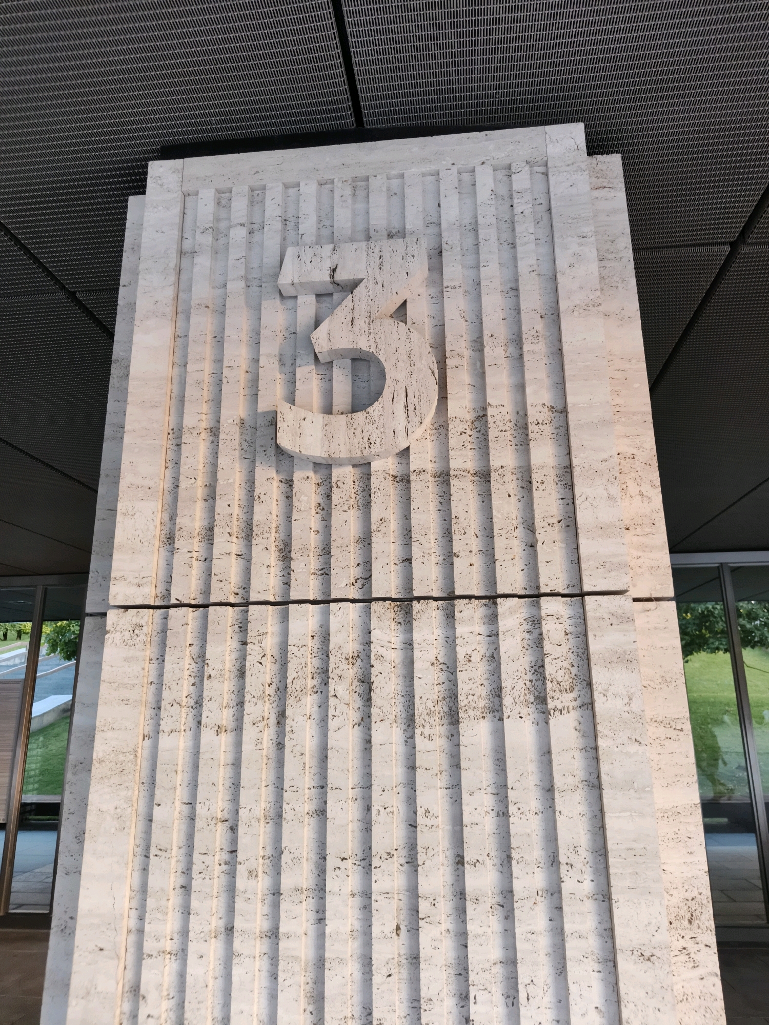 A large, textured, light-colored stone pillar stands vertically, featuring a prominent, carved numeral "3" near its top. The pillar has a fluted design with distinct vertical grooves. Above the pillar, a dark, gridded ceiling is visible, suggesting an indoor or covered outdoor space. To the sides and bottom of the pillar, reflections of greenery and glass windows can be seen, indicating the presence of an adjacent building or park. The lighting suggests it is daytime, with sunlight casting shadows on the pillar's grooves. There are no people or animals in the frame. This structure appears to be an architectural feature, possibly marking an entrance or a specific building number. Based on the available information, this pillar is located at 3 Teatralnaya Street in Krasnodar, Russia.