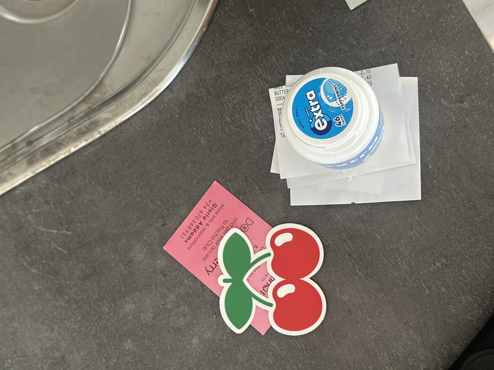 The image displays an overhead view of various items resting on a dark grey, textured surface, likely a countertop, with a metallic, possibly stainless steel, edge visible on the left. The scene suggests an indoor setting in Eivissa, Spain, a detail corroborated by text on one of the objects.
A white, circular container of "Extra" peppermint chewing gum is prominently featured, labeled with "46 PIECES" and the identifying number "5077972 2334-9". Beneath the gum container, several white paper slips, likely receipts, are stacked. Partially legible text on these slips includes numbers such as "2,10", ".40", ".15", and words like "BUTTER", "0004", and "Ta".
To the lower left, a distinct red and green cherry-shaped cutout or sticker with a white border lies atop a pink card. This card, likely promotional, provides several key details: "Do & get your access to Pacha Club", "More info & reservations", "Gloria Addams", and the Spanish phone number "+34 670568951". The words "IBIZA" and a time reference "13 PM to 1" are also visible, explicitly connecting the items to nightlife and leisure activities in Ibiza. Other partially visible text includes "erry" (likely part of a word like 'Cherry') and "anot".
No individuals are present, and no specific activity is occurring beyond the static arrangement of these everyday items. The collection, particularly the Pacha Club card and the location cue "IBIZA," strongly points to recent or upcoming leisure activities in Eivissa.