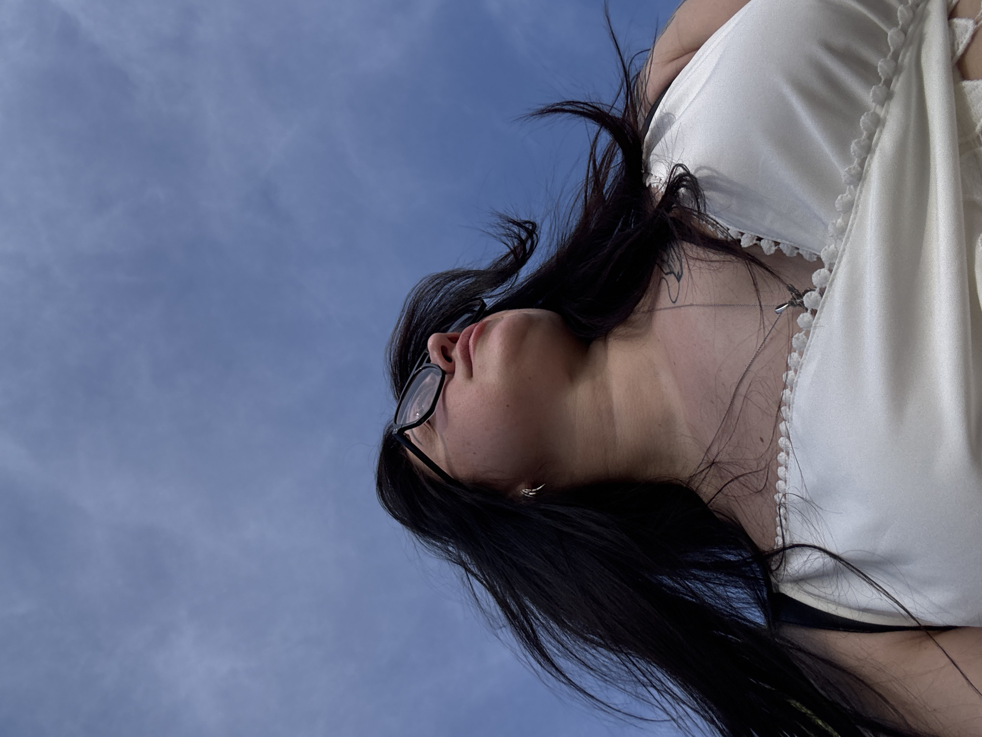 A low-angle shot features a person with dark hair and glasses looking up against a blue sky. The person is wearing a white top with pom-pom trim. The location and city are unknown.