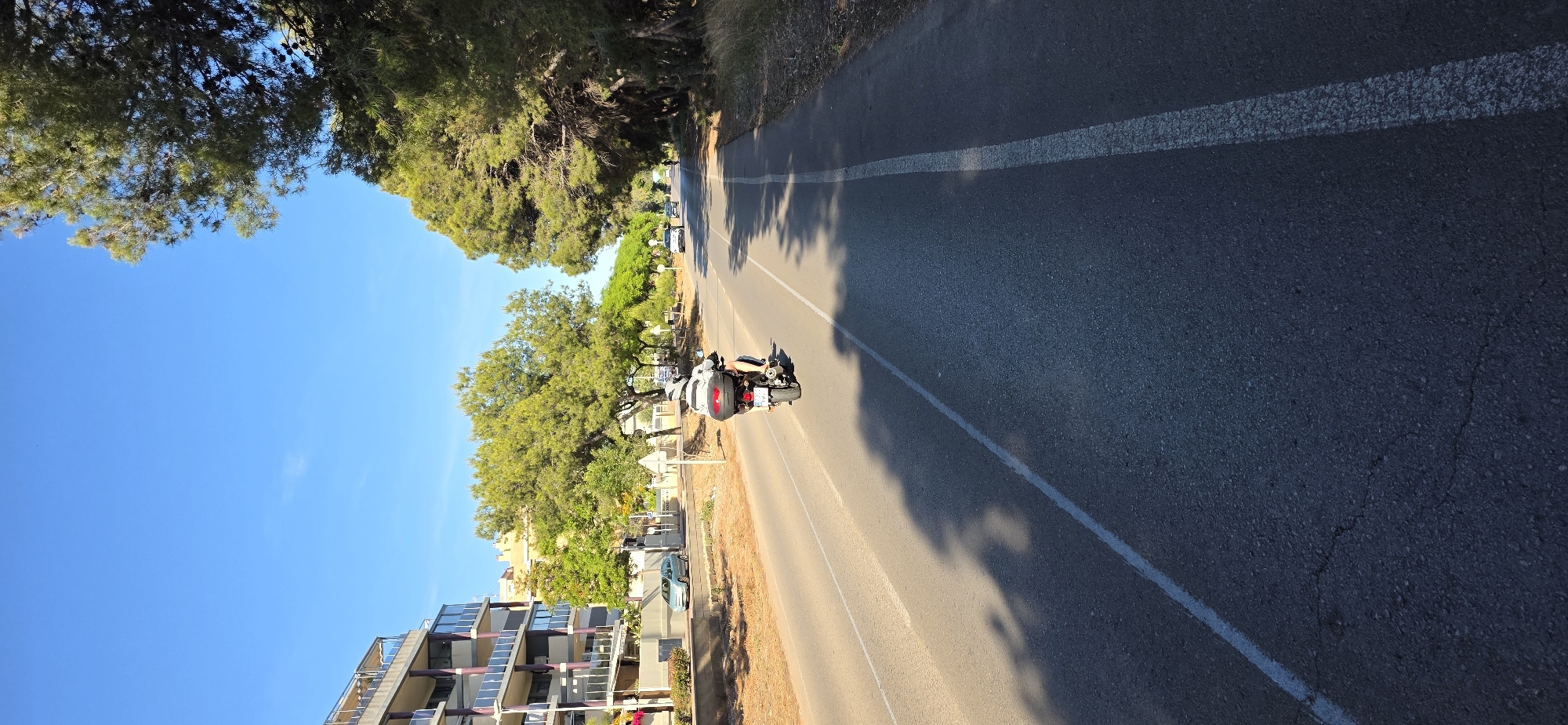 A slightly rotated, medium-angle shot captures a motorcycle moving on a road in Canet d'en Berenguer, Spain. The motorcycle is white with a single rider. The road, marked by a white line, extends into the distance with shadows cast onto its surface. To the left of the road, a multi-story residential building is visible, with balconies and solar panels. Trees line the roadside. The sky is a clear blue. Other parked cars can be seen along the roadside. The location is identifiable as Canet d'en Berenguer due to the typical architecture and landscape of the area.