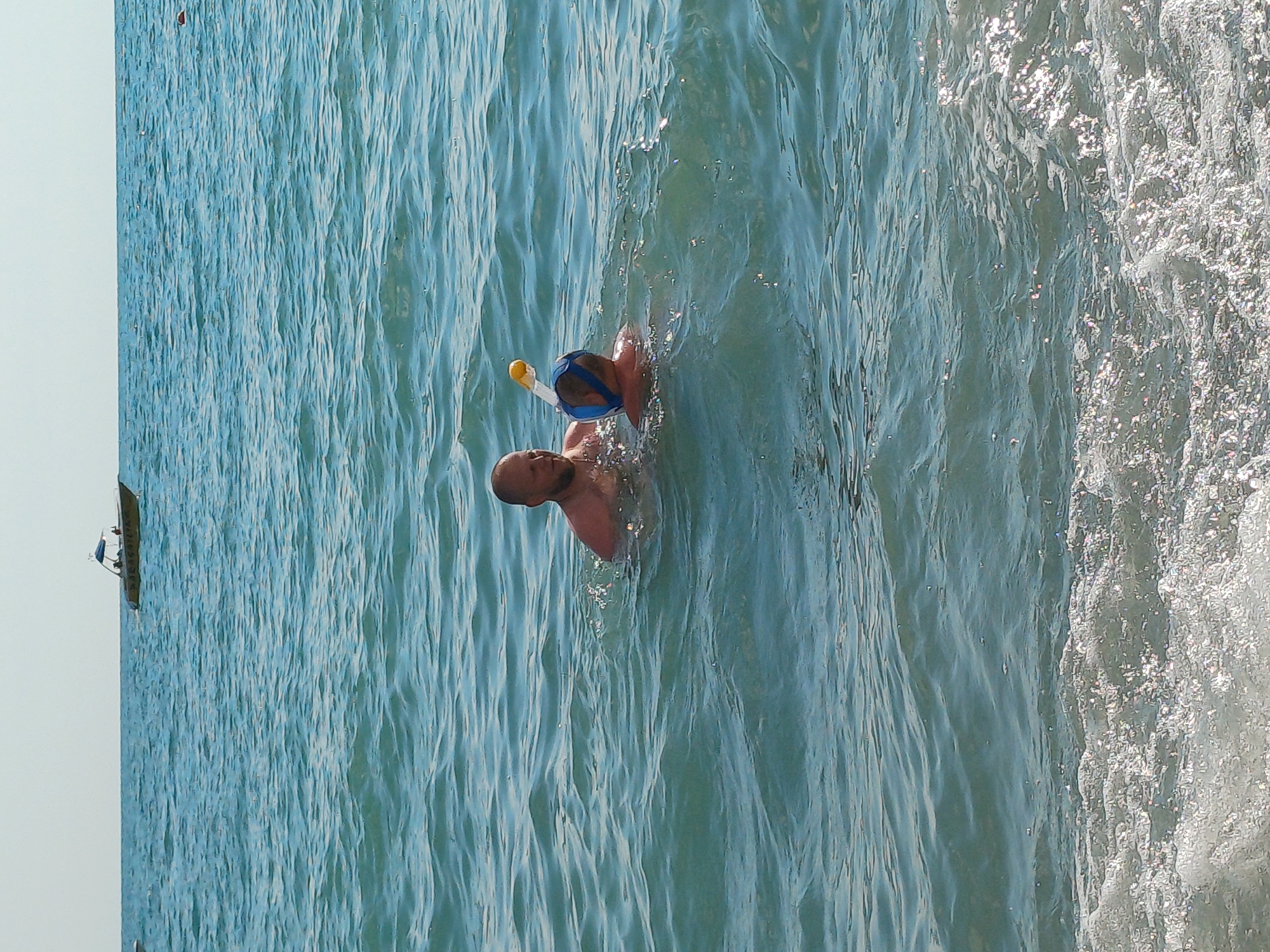 In the coastal waters of Sochi, Russia, a man and a child are swimming. The man, with a shaved head and a beard, is partially submerged in the turquoise water, his chest and shoulders visible. Next to him, the child wears a blue snorkel mask and a blue swim cap, with the yellow snorkel tube extending above the water. They appear to be engaged in swimming or snorkeling. In the background, a boat with a canopy is visible on the horizon, navigating the vast expanse of the sea under a clear sky. The water is slightly choppy, with small waves creating a sparkling effect under the sunlight.