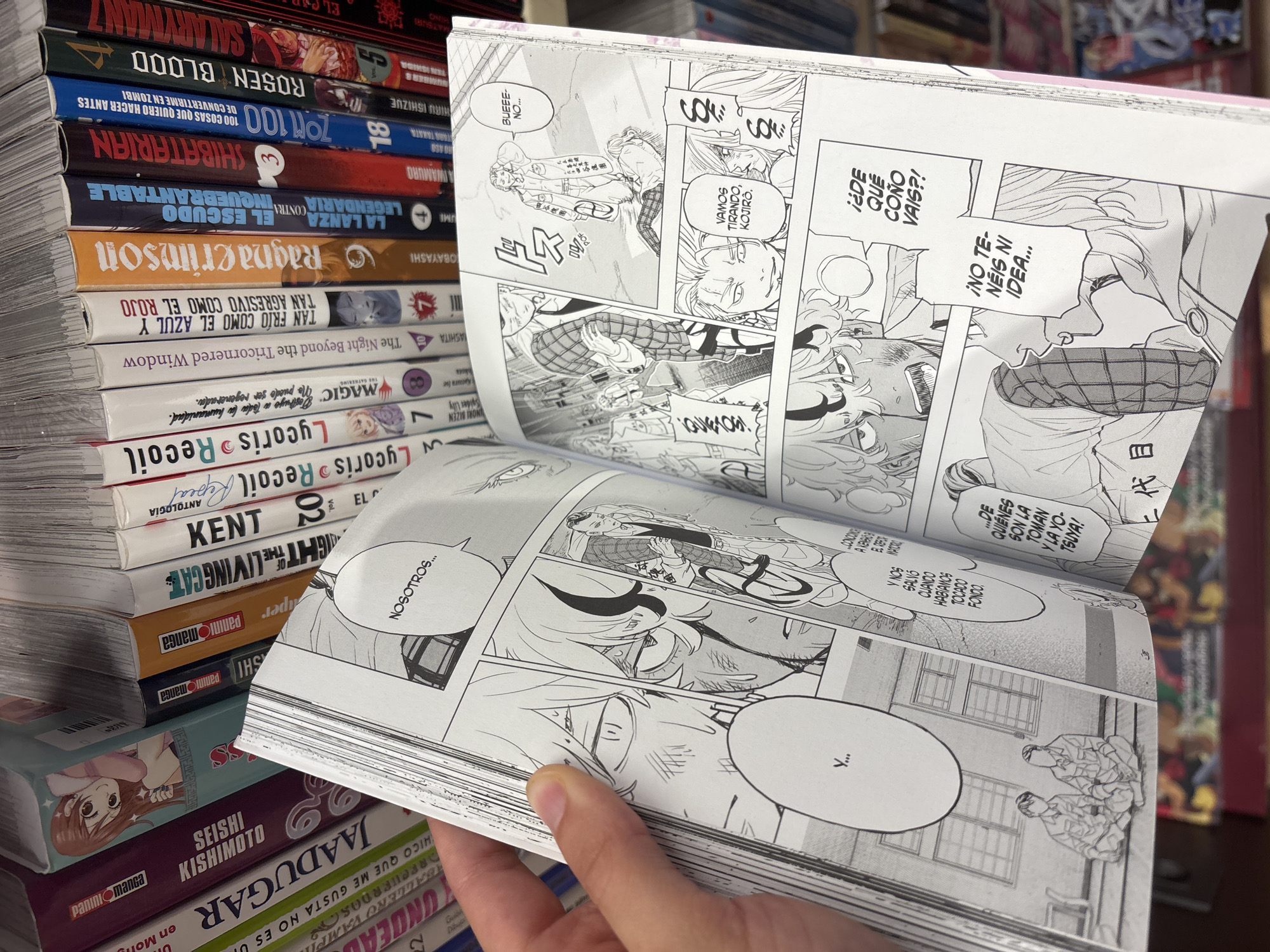 The image shows a person's hand holding open a manga book. The manga book has black and white illustrations and text. The background shows a shelf filled with various manga books, with spines of different colors and titles. The book itself is open to a page with multiple panels depicting characters in a dynamic style, with speech bubbles containing text. The environment appears to be a bookstore or library given the context of the stacked books. There is no visible indication of the city of Orihuela, Spain.