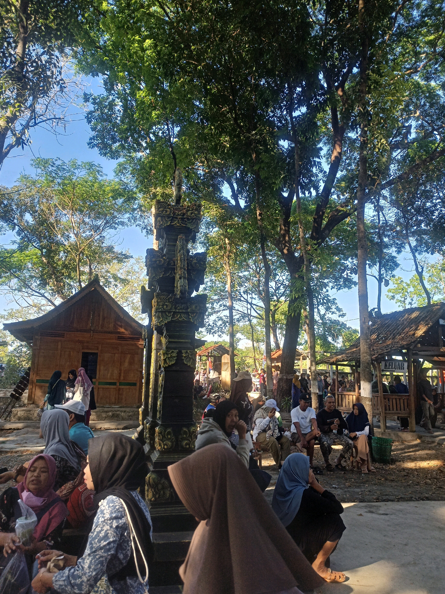 In Karungan, Indonesia, an outdoor scene unfolds under a canopy of mature trees, suggesting a natural or park-like setting. The foreground is occupied by a group of individuals, predominantly women dressed in hijabs, seated on a paved area and steps. Their postures suggest they are resting or waiting. One woman in a blue and white patterned top and black hijab is in the center, holding a small item in her hands and looking to her left. To her left, another woman in a pink and burgundy hijab is holding a plastic bag. In the background, a traditional wooden building with a peaked roof stands on a raised platform, with a few individuals visible near its entrance. To the right, a rustic open-sided shelter with a tiled roof is present, a sign with "KELUAR" (meaning "Exit" in Indonesian) is visible on its side, and several people are seated inside or nearby. A tall, intricately carved stone pillar stands prominently in the mid-ground, between the wooden building and the shelter. The ground is covered with fallen leaves and dirt, and the overall lighting indicates it is daytime, with sunlight filtering through the trees.