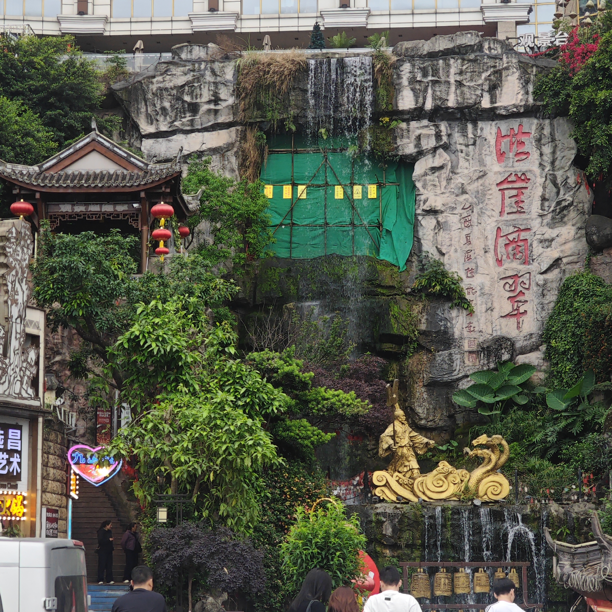 The image shows a tiered rock formation with landscaping in the Yuzhong District of Chongqing, China. At the top of the formation, a small waterfall cascades down, partially obscured by green construction netting. A golden statue of a figure and a three-headed serpent is positioned mid-way down the formation. At the base, a row of bells hang. Chinese characters are carved into the rock face. Lush vegetation is integrated into the scene. In the foreground, several people are walking or standing. To the left, a traditional Chinese-style structure with red lanterns is visible. A neon sign in the shape of a heart is lit up on a building to the left. Above the rock formation, a modern building is visible.