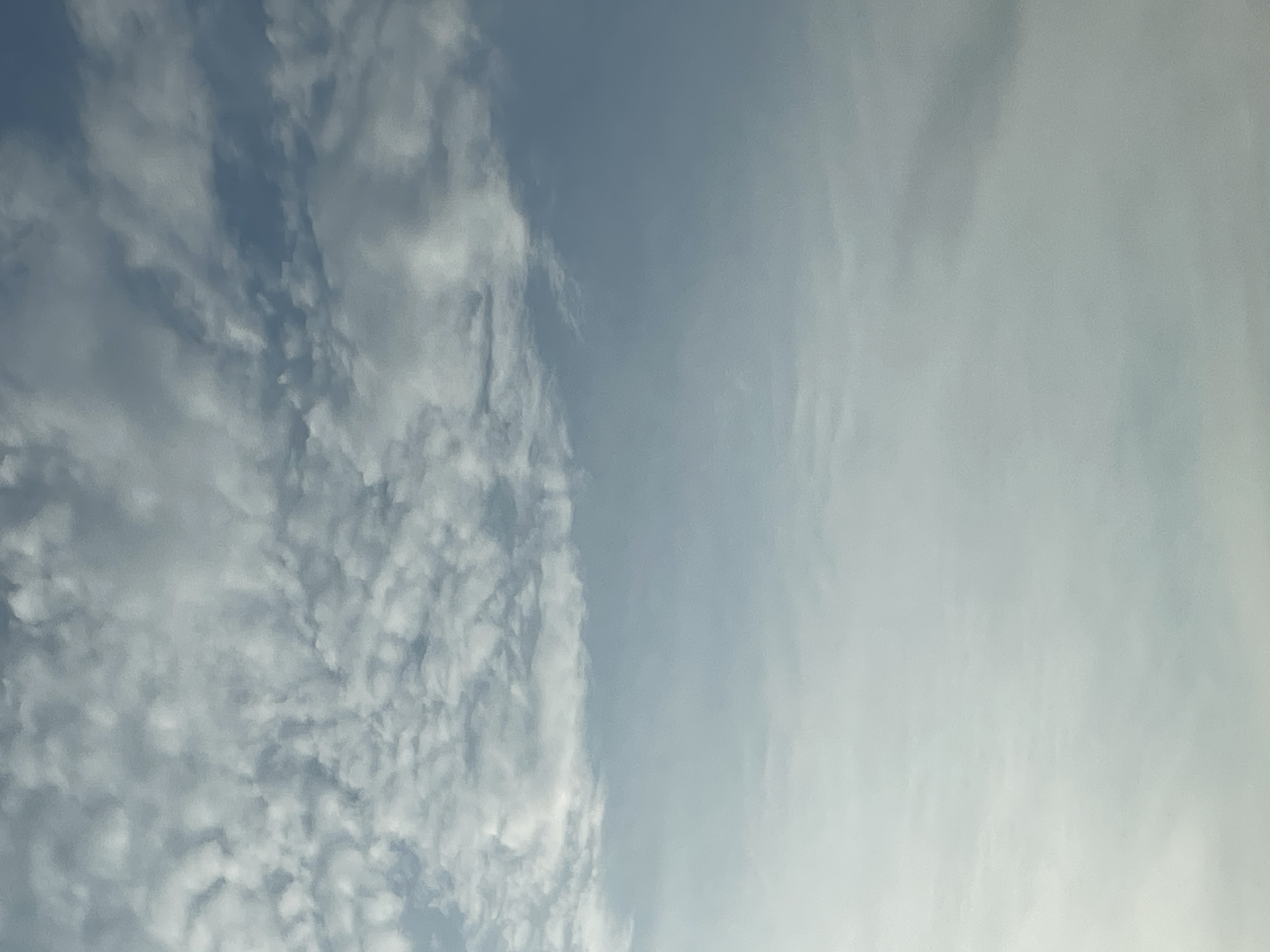 The image displays a sky filled with scattered clouds. The upper portion of the frame features denser, textured clouds, appearing whiter and more defined against the blue sky. As the view moves downwards, the clouds become lighter and more diffused, transitioning into a hazy, almost white expanse that dominates the lower half of the image. The sky itself is a pale, muted blue, suggesting either early morning, late afternoon, or a lightly overcast day. There are no discernible objects, people, or structures visible in the image; it is solely focused on the atmospheric conditions.