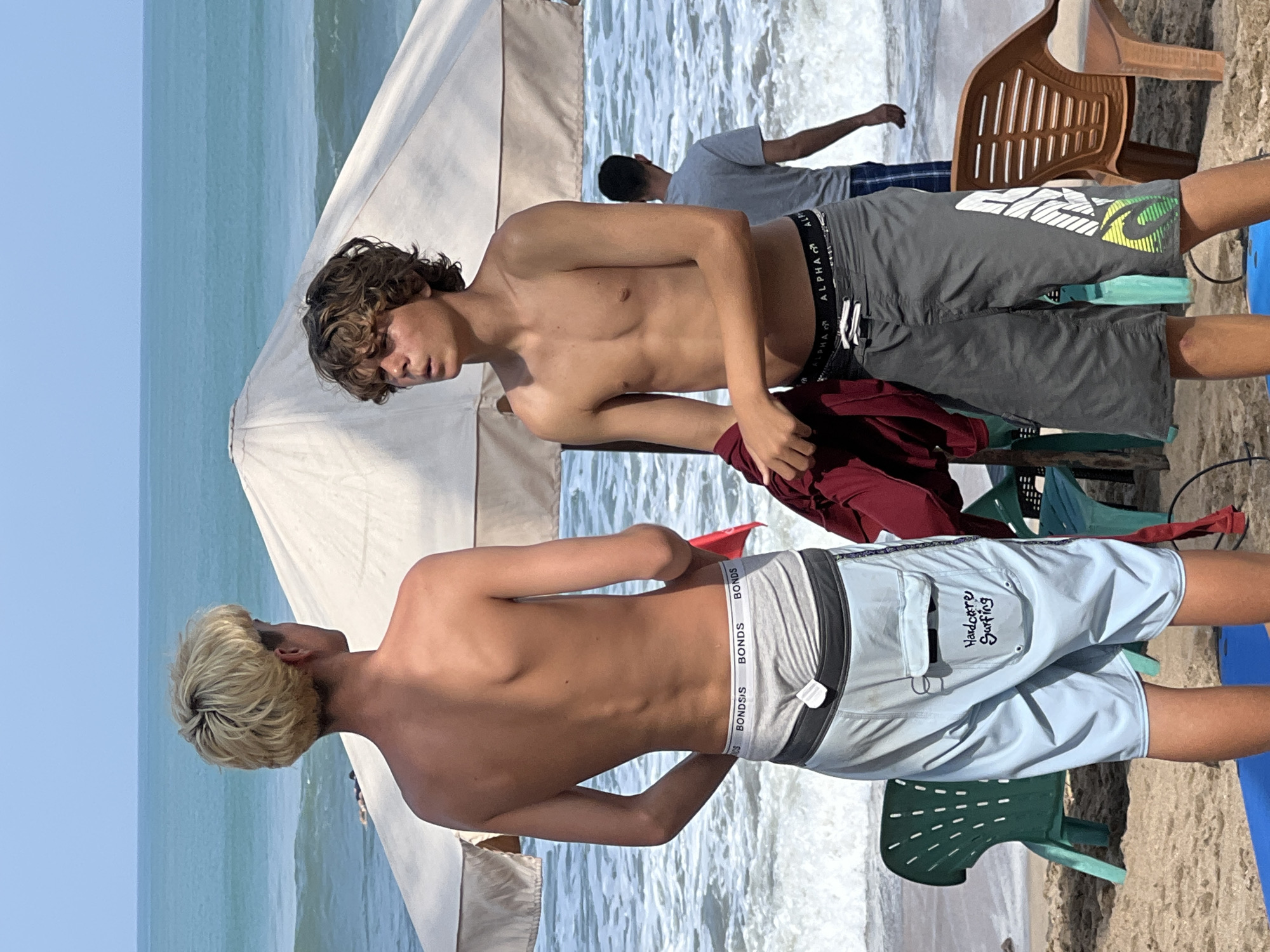 Two young men, one with blonde hair and the other with brown curly hair, stand on a sandy beach in Kuta, Indonesia, facing the ocean. Both are shirtless and wearing swim trunks. The man with blonde hair wears light blue swim trunks with a white drawstring and a grey waistband with "BONDS" repeatedly printed on it. The man with brown hair wears dark grey swim trunks with a black waistband and a white, green, and yellow graphic design on the side. The man with brown hair holds a maroon garment in his right hand. In the background, the ocean has moderate waves, and a white umbrella provides shade. A green plastic chair and a brown plastic chair are visible on the beach. Another person in a grey t-shirt stands further back, facing away from the camera. The scene appears to be during the daytime under a clear sky.