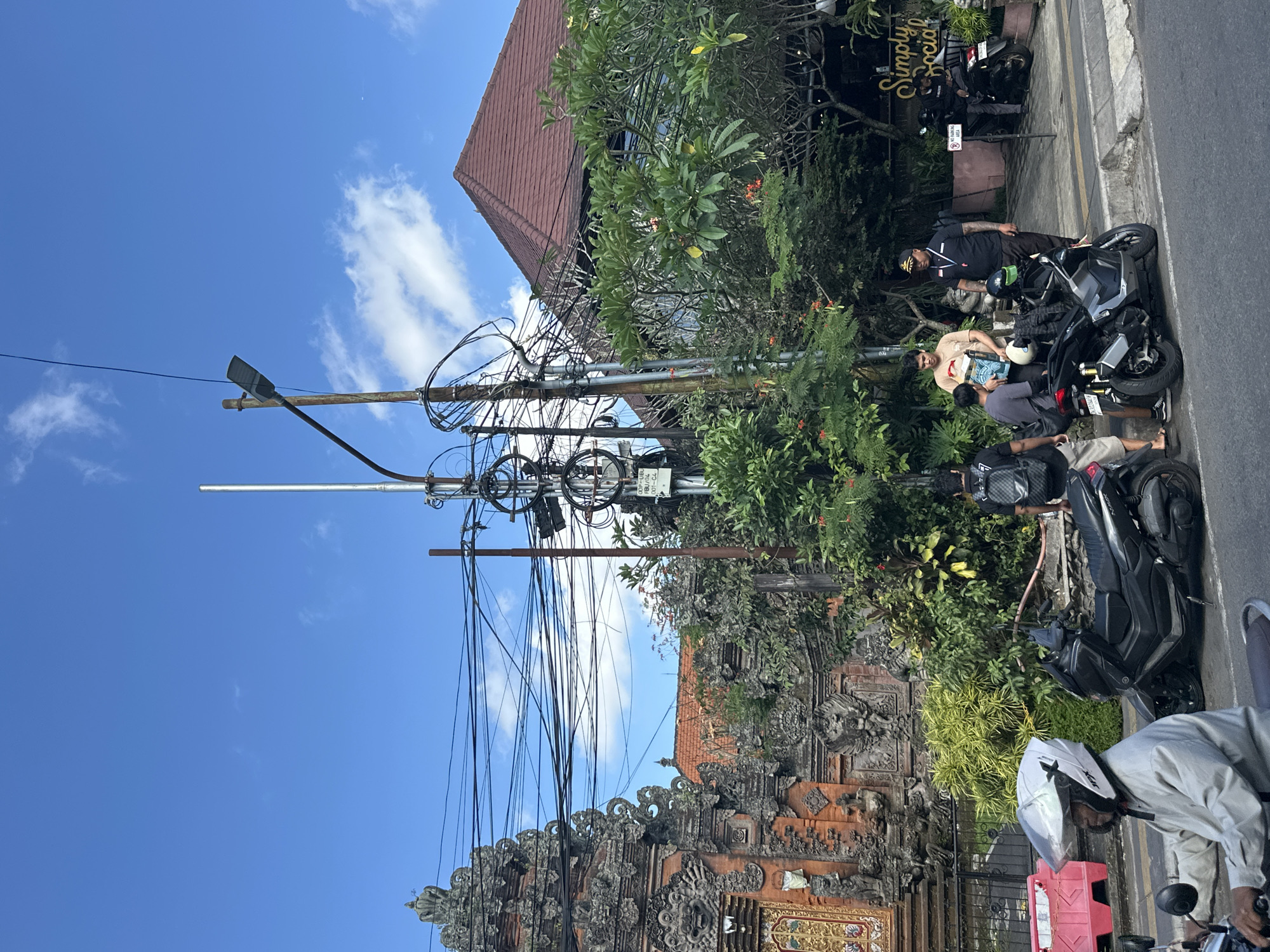 The image depicts a street scene in Ubud District, Indonesia, characterized by a mix of traditional Balinese architecture and modern infrastructure. On the left, a detailed orange stone temple gate with intricate carvings is visible. A cluster of utility poles with a dense network of black electrical wires dominates the central area, with a streetlamp attached to one of them. Green foliage and trees are abundant, with some flowering plants adding splashes of red and yellow. A red portable barrier is present on the sidewalk.
Several individuals are present. A man in a white helmet and light-colored jacket rides a motorcycle in the foreground. Behind him, on the sidewalk, a group of people are gathered. One man is carrying a shopping bag. Another man, wearing a black t-shirt and a cap, stands near a black motorcycle. Further down the street, other individuals are seen near a building with a sign that reads "Simply Social". Two black motorcycles are parked on the sidewalk. The sky is blue with scattered white clouds, suggesting a daytime scene. The buildings show red tile roofs.