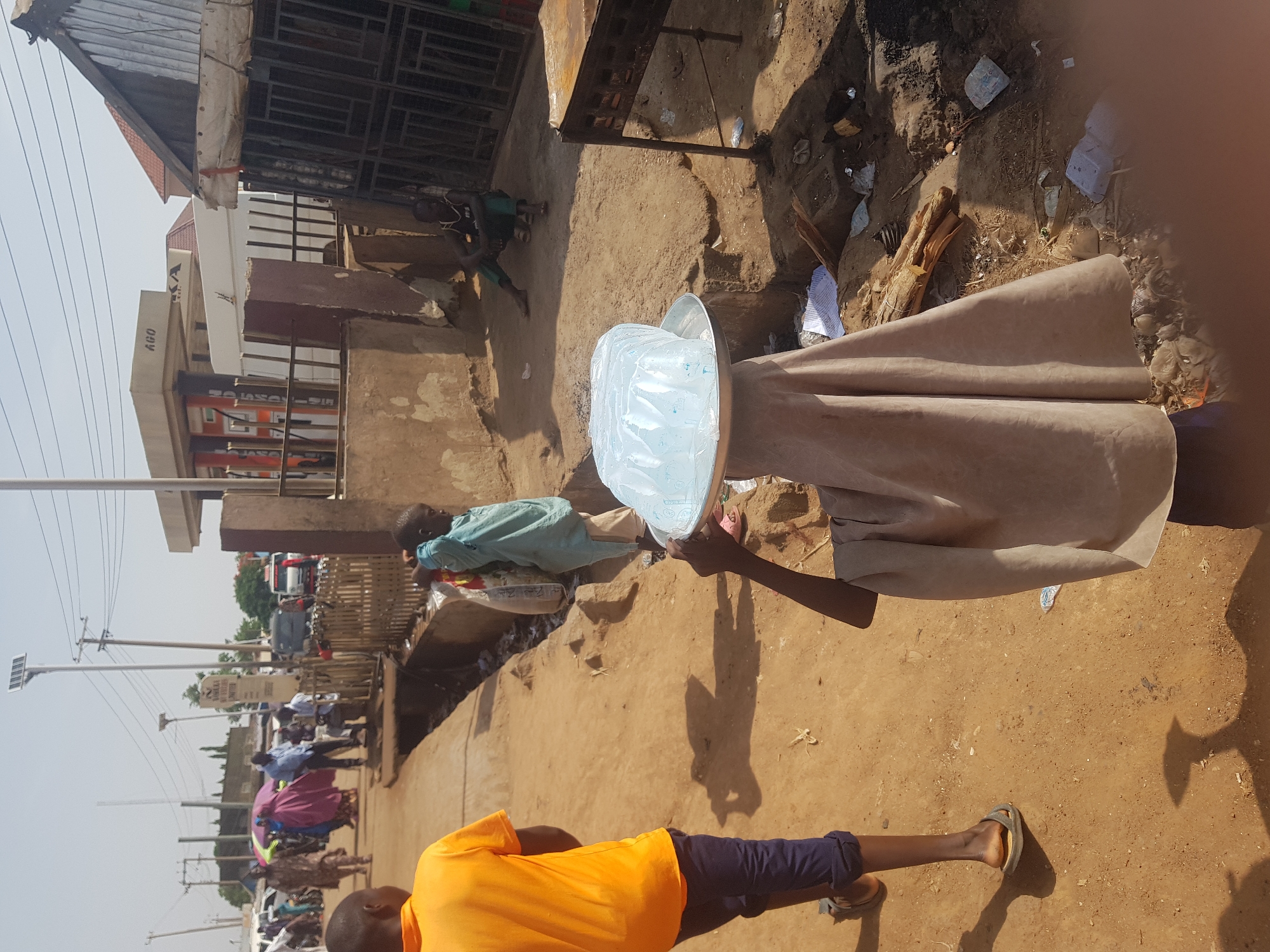 This vertical image depicts a bustling street scene in Tukur Tukur, Nigeria, under bright daylight.
In the immediate foreground, a person, mostly covered by a long, light brown garment, carries a large metal tray filled with numerous clear plastic bags of water on their head. Their right arm is visible, holding a stick. To the left of this central figure, another person in a bright orange t-shirt and dark trousers walks away from the camera. A young boy in a light blue shirt stands nearby, facing the water carrier.
The ground is unpaved, consisting of dry, light brown earth, uneven in texture, with some scattered debris and waste visible. In the background, a busy street stretches into the distance, lined with various buildings and structures, some with corrugated iron roofs. Several people are visible walking or standing along the street. Text visible on a building includes "AGO" and part of "KA". Power lines traverse the scene, and a solar-powered streetlight can be seen. Vehicles, including a truck, are present further down the road. The strong, direct sunlight creates sharp shadows, indicating a sunny midday or early afternoon.