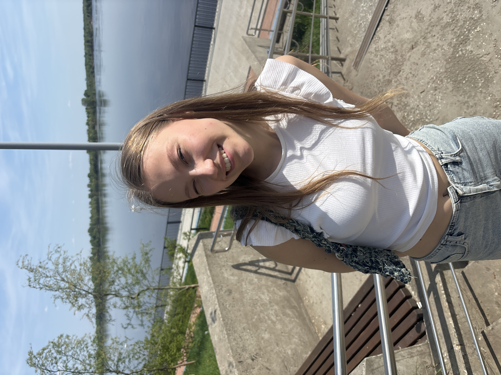 A young woman is standing on a paved waterfront area in Perm, Russia, smiling at the camera. The woman is wearing a white t-shirt and light blue jeans. She is leaning against a metal railing. In the background is a body of water and trees along the shore. The day appears sunny, casting shadows on the concrete structure.