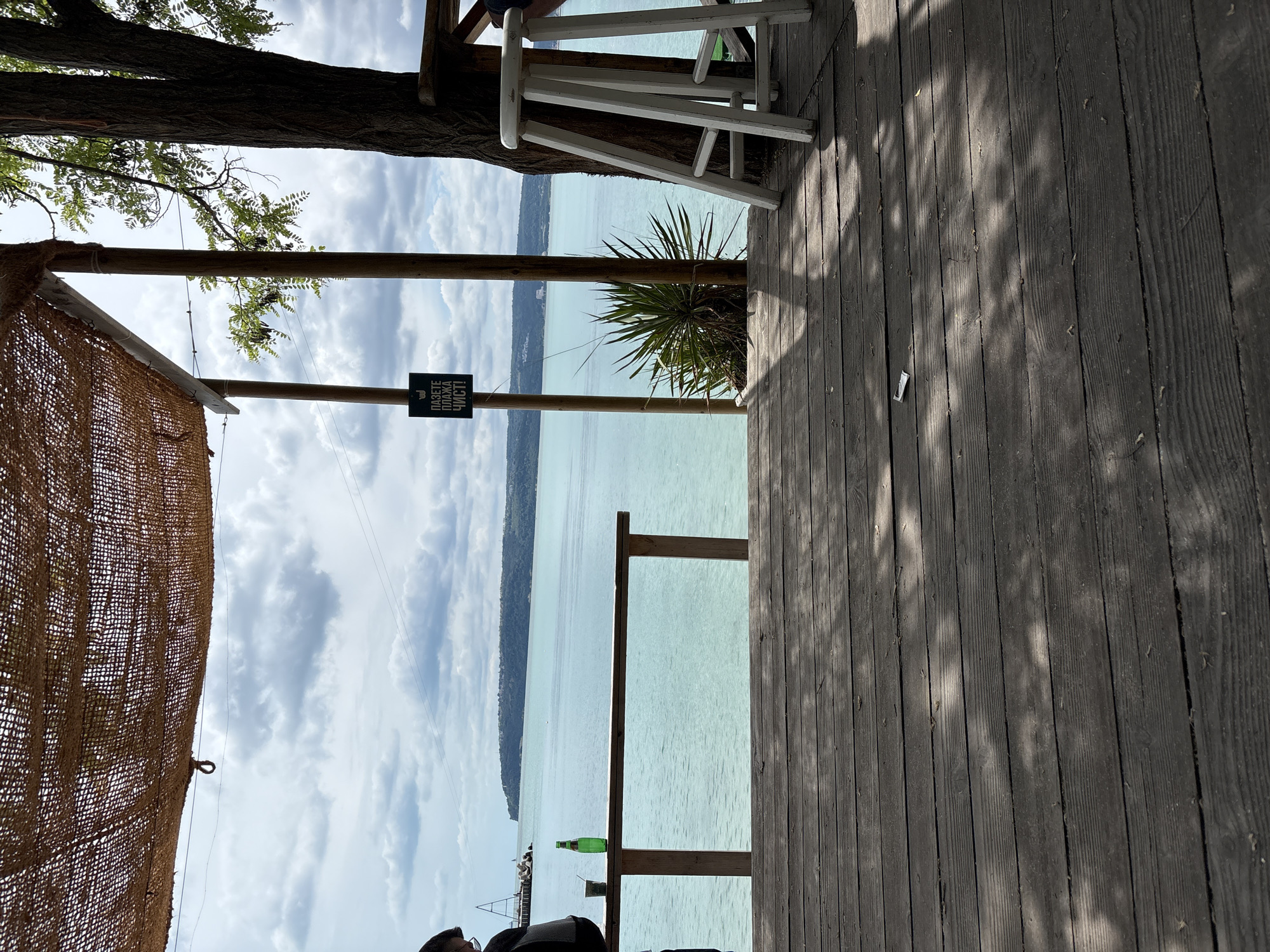 The image depicts a view from a wooden deck overlooking a tranquil, turquoise sea under a cloudy sky. A thatched roof overhead provides shade, supported by wooden posts. To the left, a person wearing a dark jacket and glasses stands, holding a phone. On a wooden railing, a green bottle and a small cup are placed. A sign with Bulgarian text, reading "PAZETE PLAZHA CHIST!" (Keep the beach clean!), hangs from a post. To the right, a white wooden stool is positioned next to a large tree, its branches casting dappled shadows on the deck. In the distance, a landmass with buildings is visible on the horizon. The scene appears to be at a beach bar or cafe in Varna, Bulgaria, specifically near the Sea Garden area, given the typical amenities and the view of the coastline. The wooden deck is aged and weathered, with scattered leaves and shadows. The overall atmosphere is relaxed and summery.