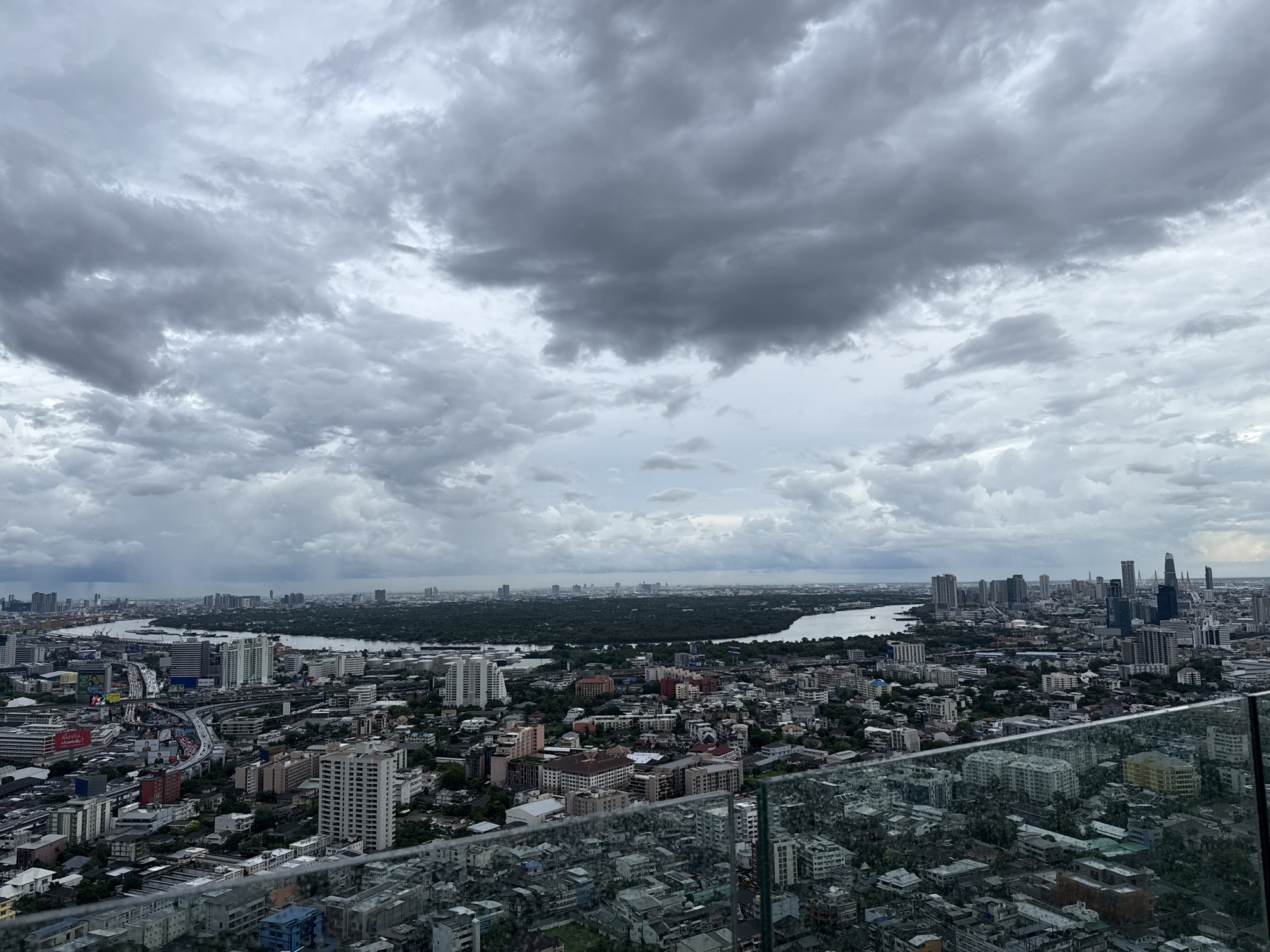 This is an aerial, wide-angle photograph of Bangkok, Thailand, taken on an overcast day. The foreground shows a glass railing of a balcony, through which a dense cityscape is visible. The city is characterized by a mix of high-rise buildings, mid-rise apartment complexes, and lower-density residential areas. Streets with traffic are visible, winding through the urban landscape.
In the mid-ground, a wide river, likely the Chao Phraya River, curves through the city, bordered by lush green trees and vegetation on its banks. Several bridges cross the river, and boats can be seen on its surface. Beyond the river, the city continues with more buildings and a distant horizon.
The sky is filled with dramatic, heavy grey clouds, suggesting an impending storm or recent rainfall. The lighting is diffused and moody, casting a subdued light over the entire scene. There are no identifiable people or specific actions depicted in the image. The overall impression is a vast, sprawling metropolitan area under a stormy sky.