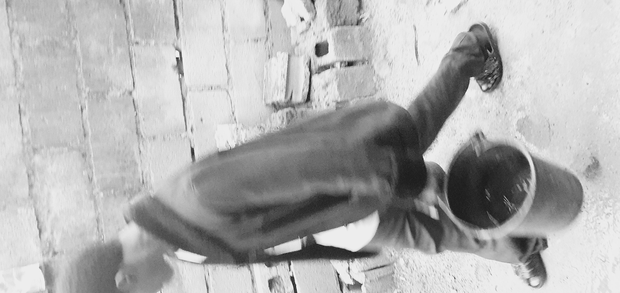 A black and white, low-resolution, motion-blurred image depicts a person walking with a bucket. The person, viewed from the side and slightly from behind, is wearing a dark jacket, dark pants, and sandals. They are carrying a dark bucket in their right hand, which appears to contain liquid. The background consists of a brick wall and scattered bricks on the ground. The scene is outdoors, with a rough, unpaved surface beneath the person's feet. The overall impression is one of movement and activity, possibly in a construction or work area. The image quality and motion blur make specific details about the individual's facial features, clothing patterns, and the exact contents of the bucket indistinct. The location is identified as Anguldi, Nigeria.