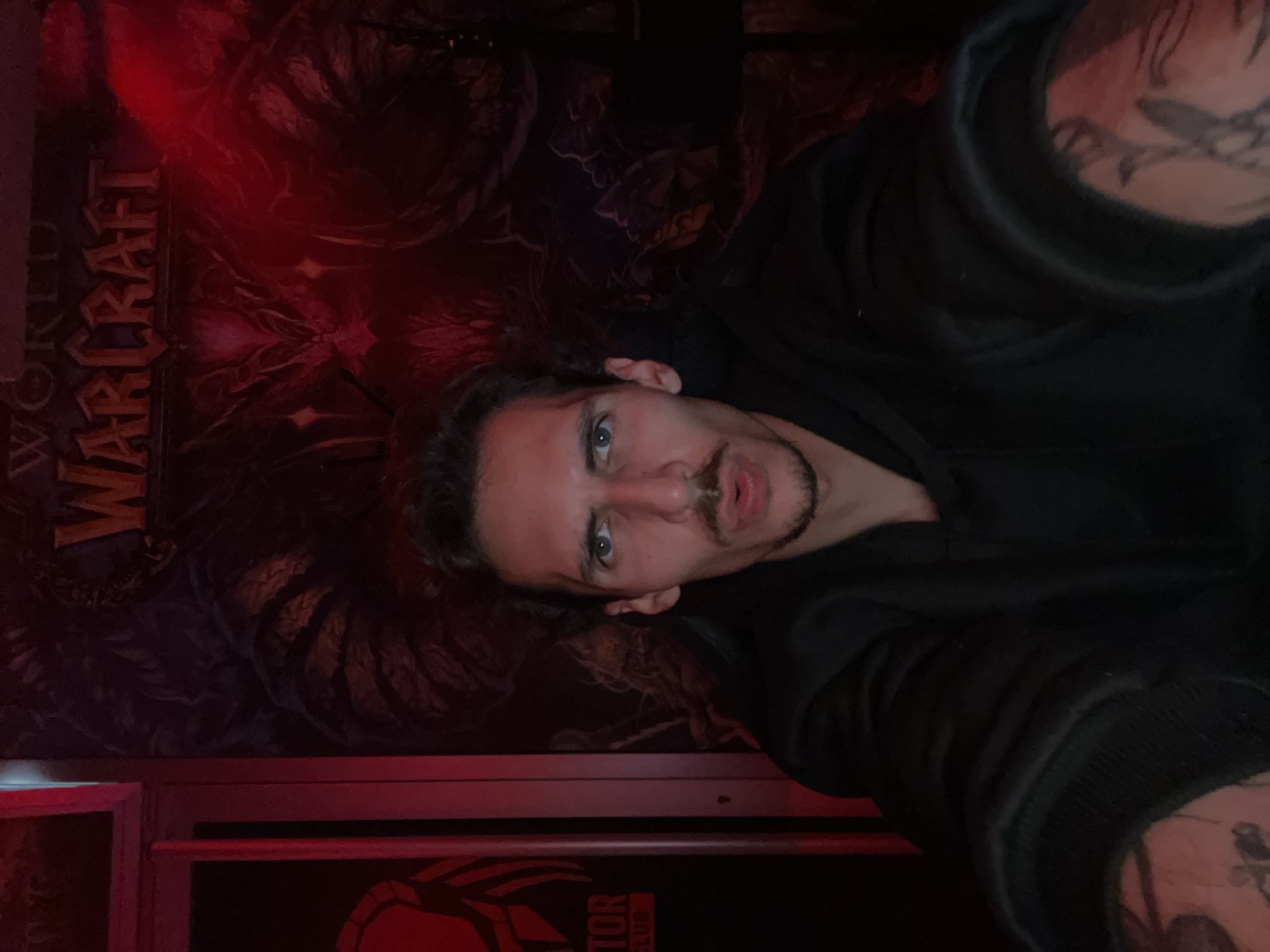 A male individual is captured in a selfie, looking directly at the camera with a neutral expression. He has dark, wavy hair, a mustache, and a goatee, and his eyes are light-colored. He is wearing a dark, possibly black, hoodie or long-sleeved top, and tattoos are visible on his left forearm and upper arm.
The setting is an indoor space characterized by dim, predominantly red lighting, which casts a strong hue over the scene. The background is dominated by a large poster or tapestry. The most prominent visible text on this poster is "WORLD WARCRAFT," displayed vertically on the left side, against a dark, intricate fantasy-themed artwork depicting what appears to be monstrous or mythical figures, consistent with the game's aesthetic.
Below the large poster, at the bottom of the image, a partial red logo or text is visible on a dark surface. The letters "TOR" are clearly legible, followed by obscured characters, suggesting part of a word like "PREDATOR" or "VICTORIOUS," likely related to gaming or technology given the "World of Warcraft" theme.
The overall ambiance suggests a gaming-oriented environment, such as a personal gaming room or a themed establishment, possibly captured during the evening or night due to the artificial lighting. There are no specific visual cues within the image to identify the location as Budva, Montenegro, other than the provided context.
