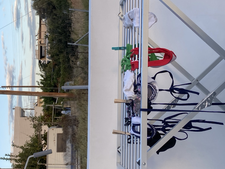 The image depicts an outdoor scene in Skala, Greece. In the immediate foreground, a white-painted parapet or wall is present. Directly behind and elevated above this parapet is an aluminum-colored, collapsible clothes drying rack. Numerous items of wet laundry are drying on the rack, including multiple dark blue and white striped bikini tops and bottoms, a plain dark blue bikini bottom, red garments, green garments, white socks, and a white folded garment. These items are secured by several wooden clothespins and one green plastic clothespin. An angled grey metallic structure is partially visible on the far left of the rack.
The midground reveals a transitional area behind the drying rack and parapet. This area consists of overgrown dry grass and scattered green vegetation. A metal post and wire fence runs horizontally through this section. A white caravan or mobile home is parked on the left side, partially obscured by foliage. Beyond the caravan, a portion of a white building with a flat roof is visible. A tall, dark brown wooden utility pole with attached wires stands prominently to the right of the caravan. Another thinner grey pole is visible closer to the foreground.
In the background, typical residential buildings with white walls and flat roofs are visible, interspersed with green trees and bushes. Some buildings feature visible solar water heaters or satellite dishes on their roofs. The sky above is partly cloudy, displaying patches of blue sky and white cumulus clouds, indicating daylight conditions. No individuals or active interactions are depicted in the scene.