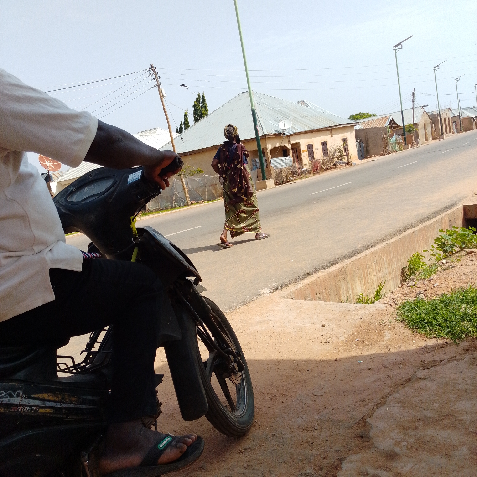 The image depicts a street scene in Gombe, Nigeria, with a person on a motorcycle in the foreground, facing away from the camera and to the left. The rider wears a white long-sleeved shirt and dark trousers, with one foot in a black sandal visible. The motorcycle is black, with part of the dashboard and handlebars in view. The rider's right hand is on the handlebar.
In the mid-ground, a woman walks along the right side of the road, also facing away from the camera and slightly to the left. She is dressed in a patterned brown and green wrapper and a dark top, with a headscarf. She is wearing sandals.
The street is paved with asphalt and has a white lane marking. Buildings line the right side of the street, appearing to be residential or commercial structures with corrugated metal roofs and some with visible windows. Several solar-powered streetlights are positioned along the road. The environment is outdoors under a clear, bright sky, suggesting a sunny day. The ground in the foreground is a mix of dirt and some concrete elements, with sparse vegetation. The overall impression is a typical street scene in a Nigerian city.
