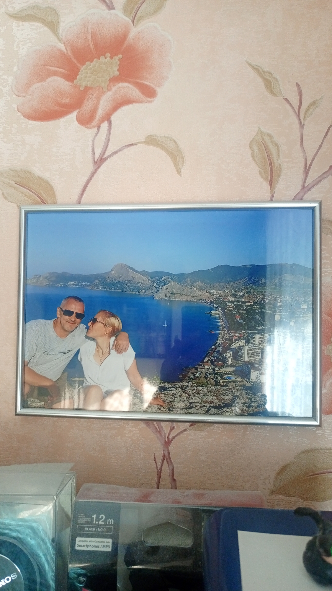 A framed photograph hangs on a wall adorned with a floral wallpaper pattern. The photograph depicts a couple sitting outdoors with a scenic coastal city and body of water in the background. The man, wearing a grey t-shirt and sunglasses, has his arm around the woman, who is wearing a white shirt and sunglasses and is leaning her head towards him. The city in the background features numerous buildings and mountains. Below the photograph, several boxes are stacked, including one with text indicating "1.2 m BLACK / NOIR" and "Smartphones / MP3". The overall setting appears to be an indoor environment. There is no visual information in the image to determine the specific location within the city of Belorechensk, Russia.