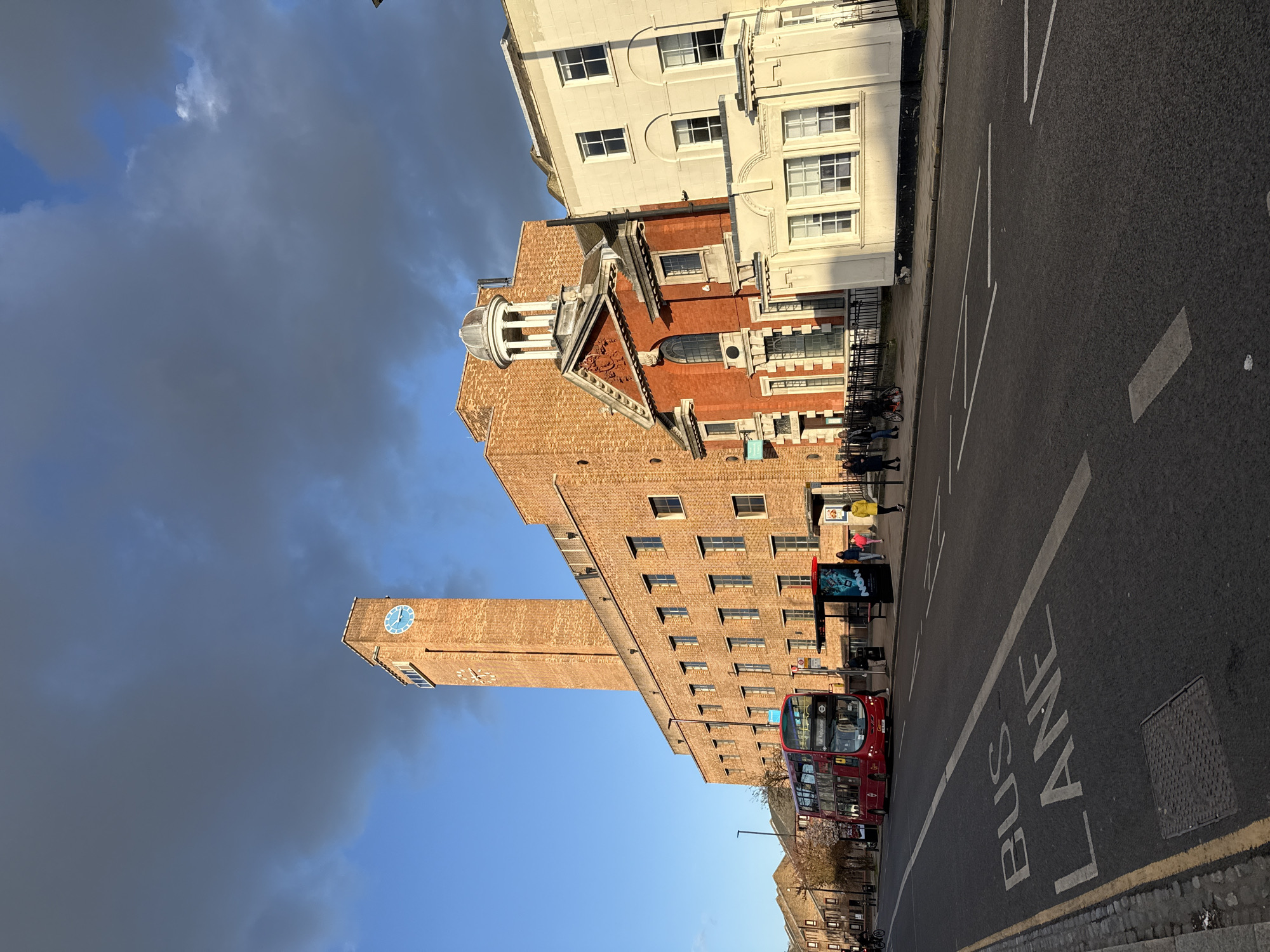 The image shows a street scene in London, United Kingdom. The main subject is a large brick building with a clock tower. A red double-decker bus is stopped in a bus lane on the right side of the street. A bus stop with an advertisement is located near the bus. Several people are standing near the bus stop. The sky above the building is partly cloudy, with dark storm clouds on the left side of the frame. There are other buildings visible on the street, including a white building next to the brick one.