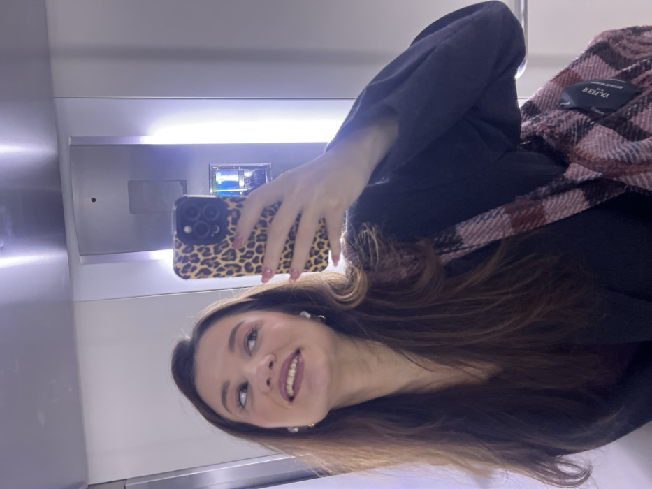 The image depicts a female individual taking a mirror selfie within an interior space, consistent with an elevator car. The specific location within Budva, Montenegro, cannot be identified from the visual context. The woman has long brown hair, a medium-light complexion, and is smiling, looking upwards and to her left. She is wearing a dark-colored jacket over a dark top, with a pink, red, and dark plaid shoulder bag, which has a "RESERVED" label visible. Her right hand, showing pink nail polish, holds a smartphone encased in a leopard print cover, angled towards the reflective surface to capture the image. Small white stud earrings are visible. In the background reflection, a light-colored panel with an elevator control panel, including an illuminated button displaying the number "1", is discernible. The environment features bright artificial lighting from overhead and vertical strips, along with light-colored, possibly metallic or laminated, wall panels.