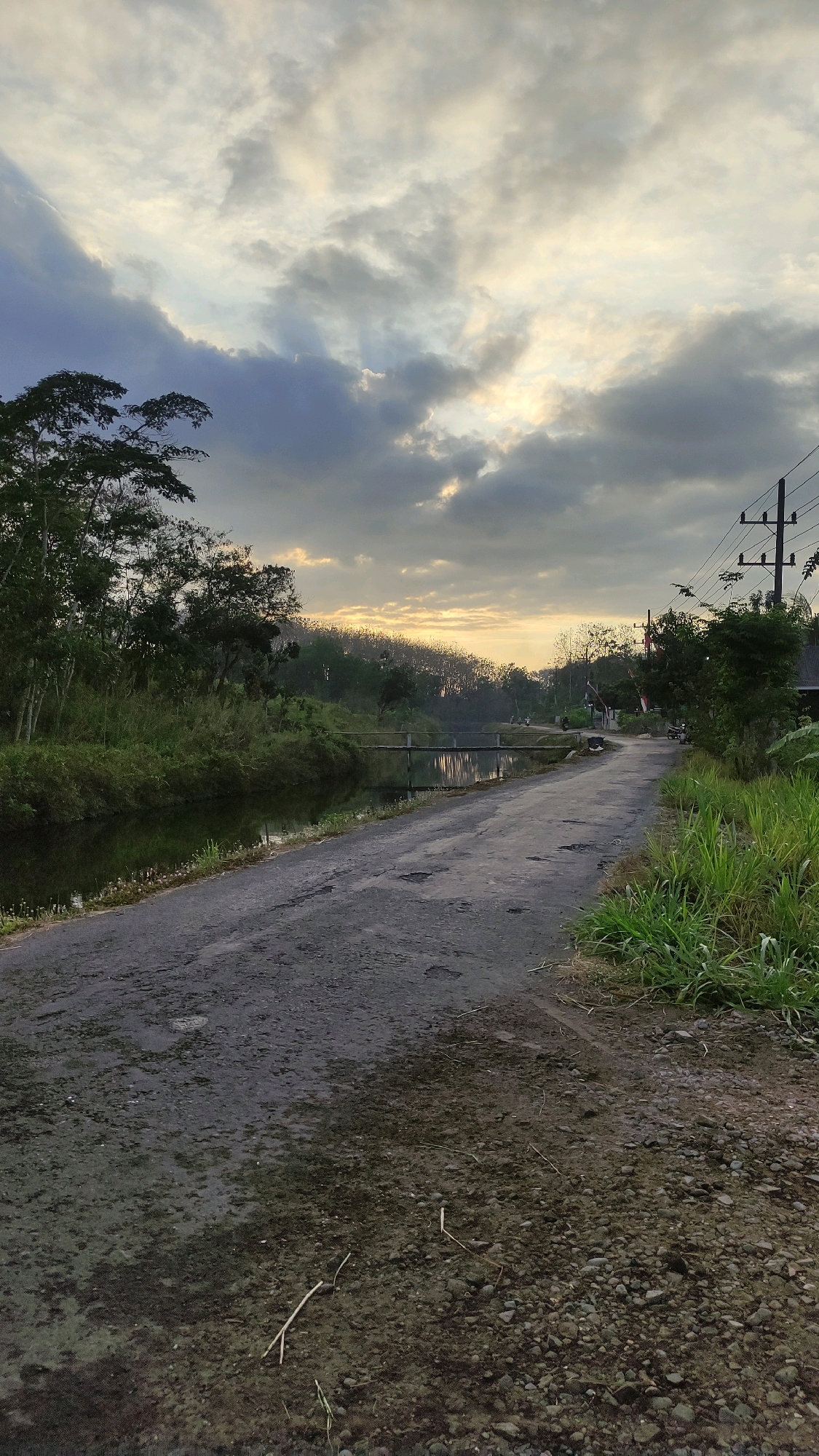 This image depicts a rural road in Tiudan, Indonesia, with a canal running alongside it. The road surface is asphalt, showing signs of wear and tear with patches and puddles. Dense green vegetation lines the canal and the right side of the road. In the distance, a simple wooden bridge crosses the canal, and a few scattered trees dot the landscape. The sky is overcast with dramatic clouds, and the sun is setting or rising, casting a warm, diffused light. There are no people visible in the scene. The overall atmosphere is serene and natural.