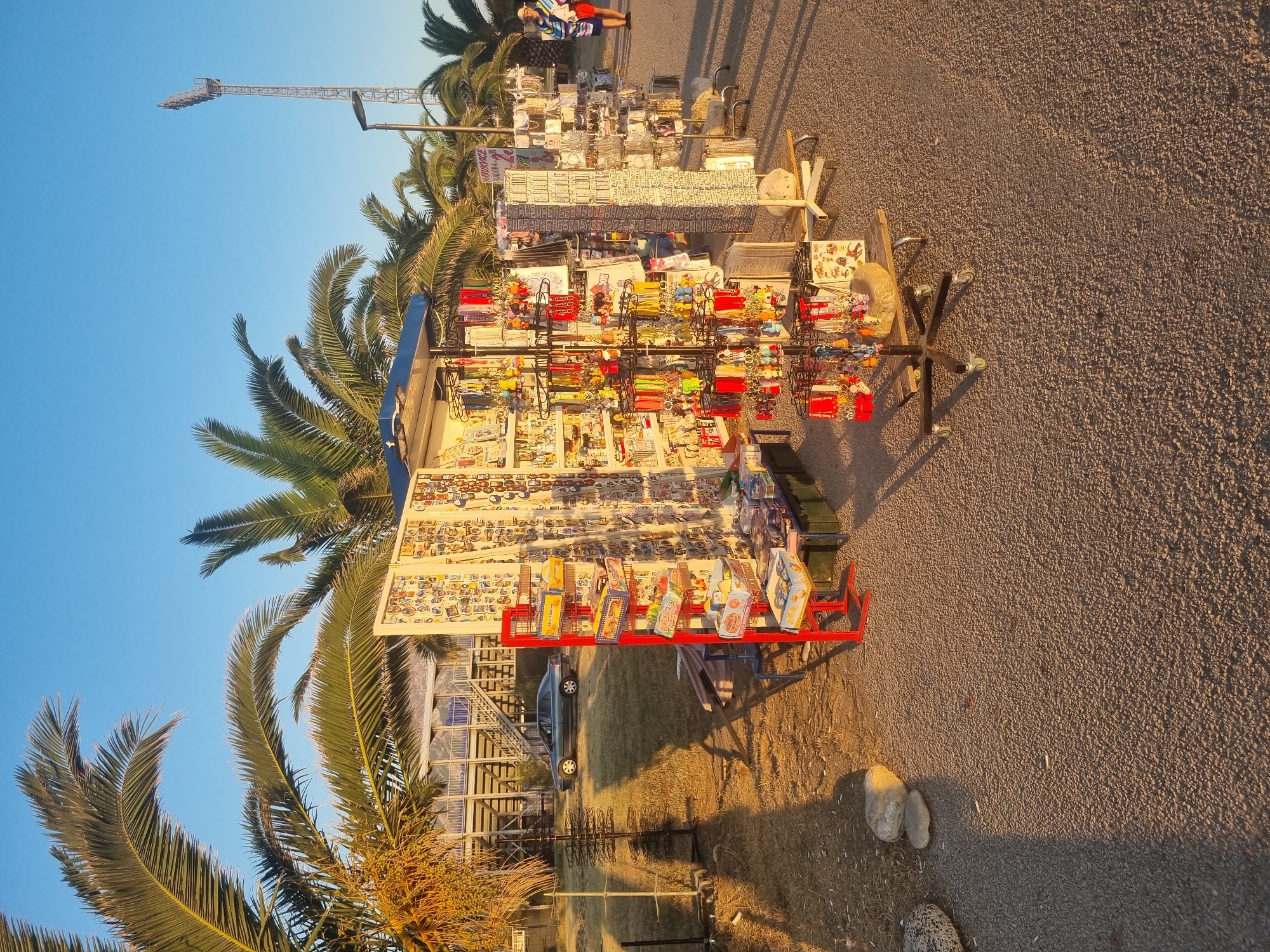The image depicts a souvenir stand set up on a paved area, likely a promenade or walkway, in Bar, Montenegro. The stand displays an assortment of trinkets, including keychains, magnets, and other small souvenirs. Palm trees are visible in the background, suggesting a coastal or warm climate. A stadium light tower can be seen in the distance. A silver car is parked on a grassy area behind the stand. A person wearing a hat and colorful clothing is walking away from the stand on the right side of the image. The sun is low in the sky, casting long shadows and illuminating the scene with a warm light. The ground is covered in a gravelly, asphalt-like material.