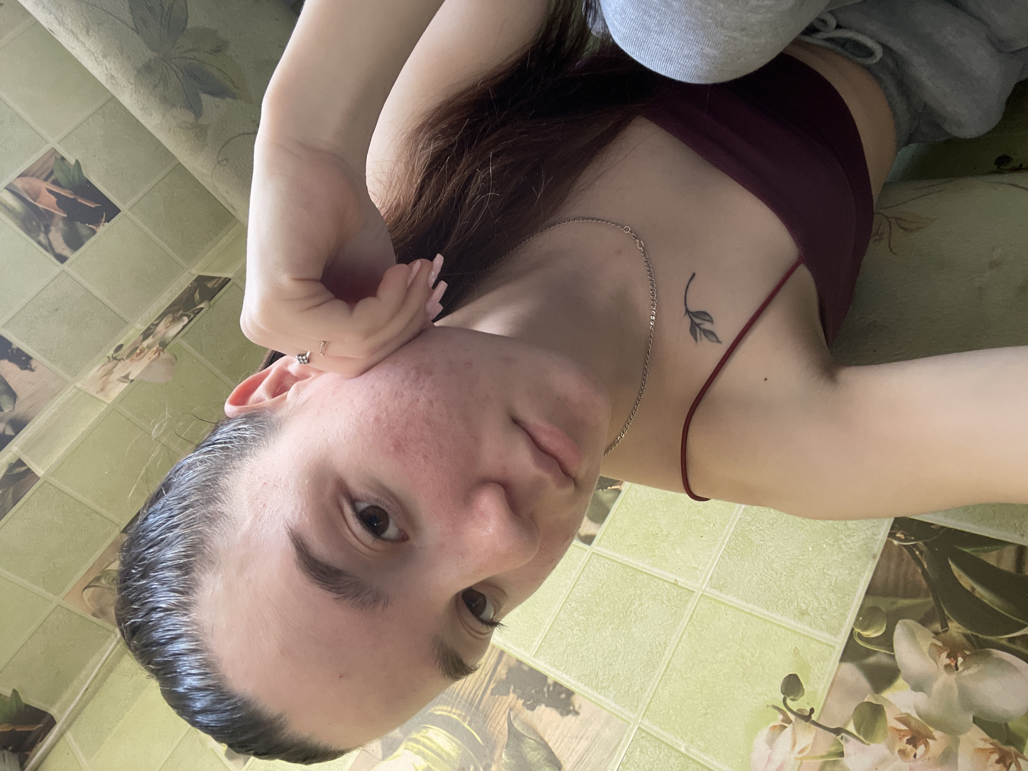 A young woman is lying down, looking at the camera. She is wearing a maroon tank top and gray sweatpants. She has long, dark hair and a small tattoo of a leaf on her chest. The background is a tiled surface. The location is unspecified and cannot be discerned from the image.