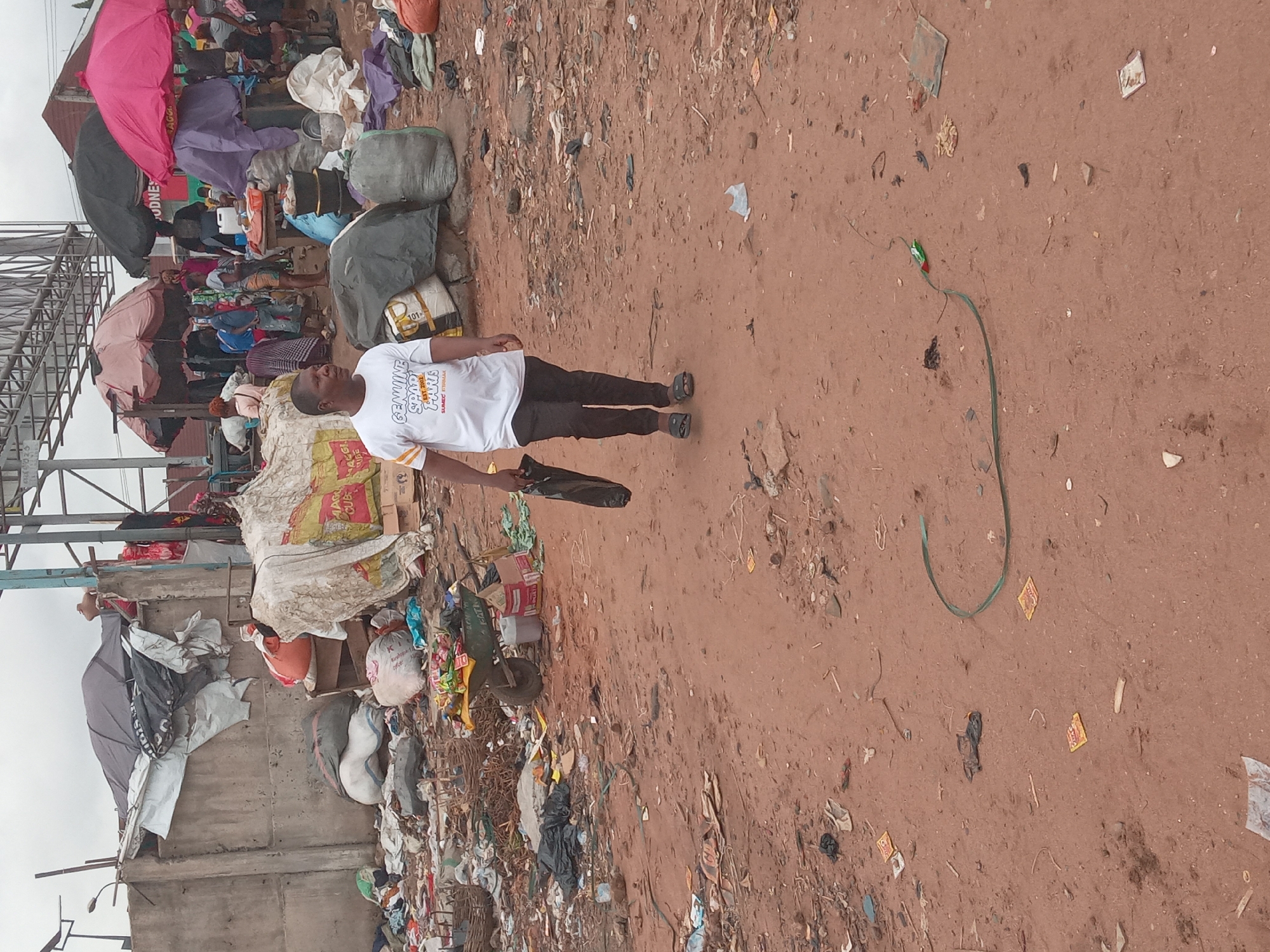 A street scene in Onitsha, Nigeria depicts a man standing amidst a littered environment. The man, wearing a white T-shirt and dark trousers, holds a closed umbrella. He stands on a dirt ground strewn with various debris and litter. To the man's left, there is a large pile of refuse including sacks, bags, packaging materials, and general trash. Behind the refuse, there are some structures, including what appears to be market stalls covered with umbrellas and other temporary roofing. In the distance, there are other people and structures, suggesting this might be a marketplace or busy street. A long, green object (possibly a hose) is lying on the ground in front of the man.